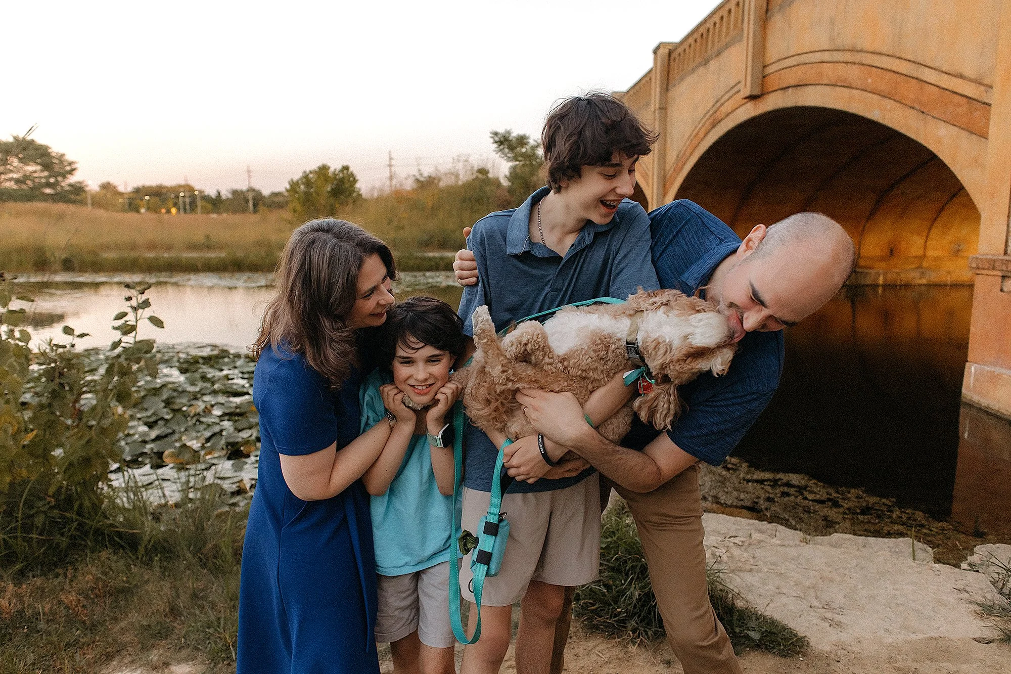 A family of four and their dog laugh by a pond near a stone bridge at sunset—captured by your Westfield Family Photographer.