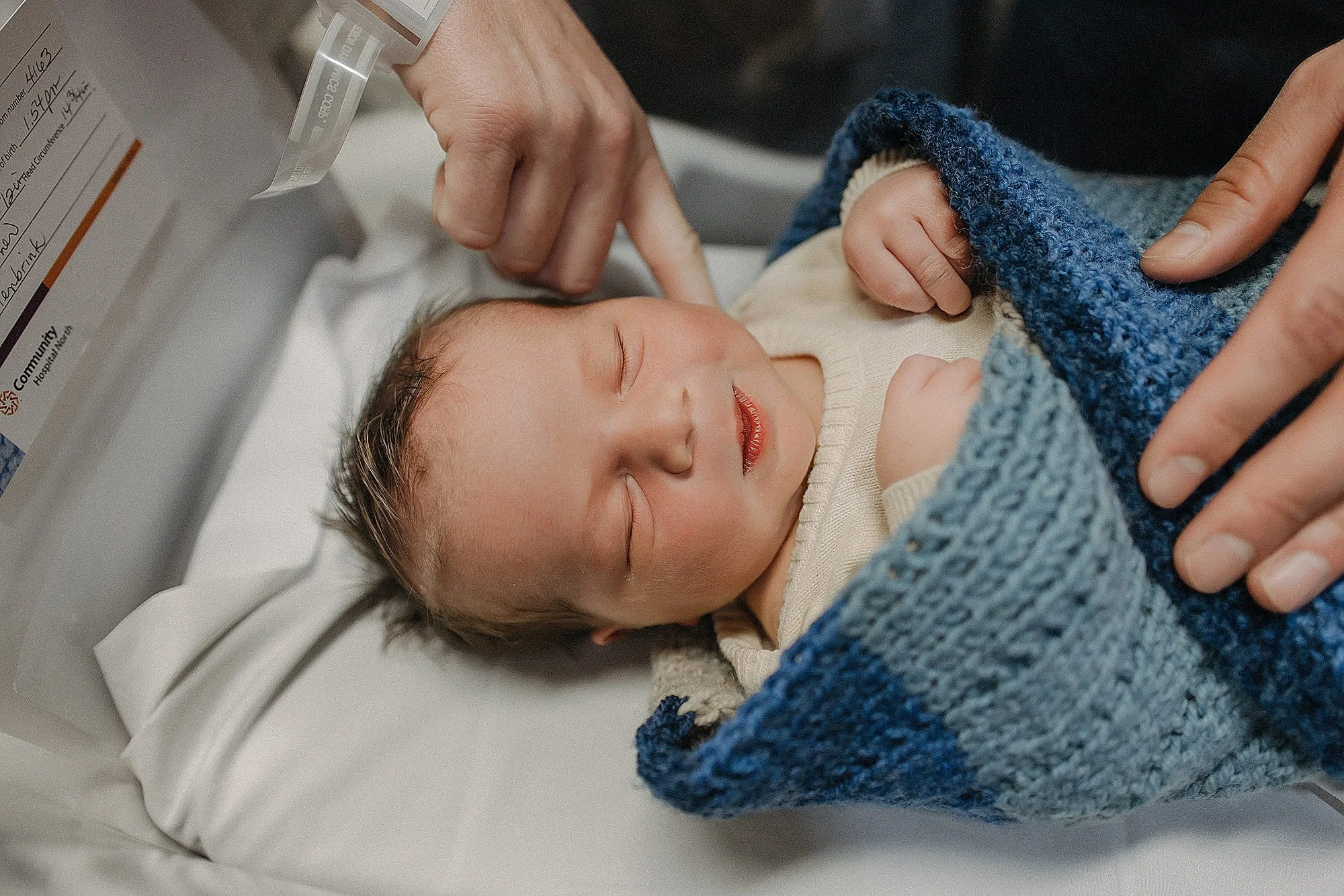 A sleeping baby wrapped in a blue and gray knitted blanket in a hospital setting.