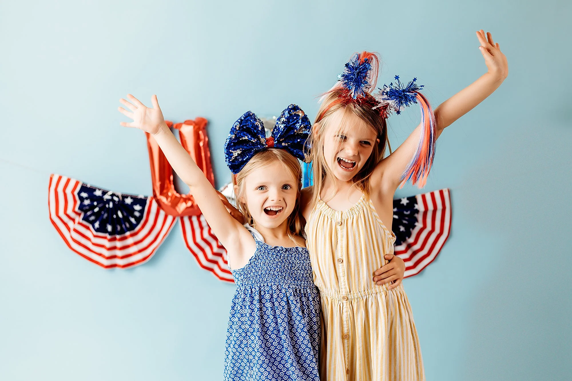 Two young girls dressed in patriotic themed outfits celebrating, with red, white, and blue party decorations in the background.