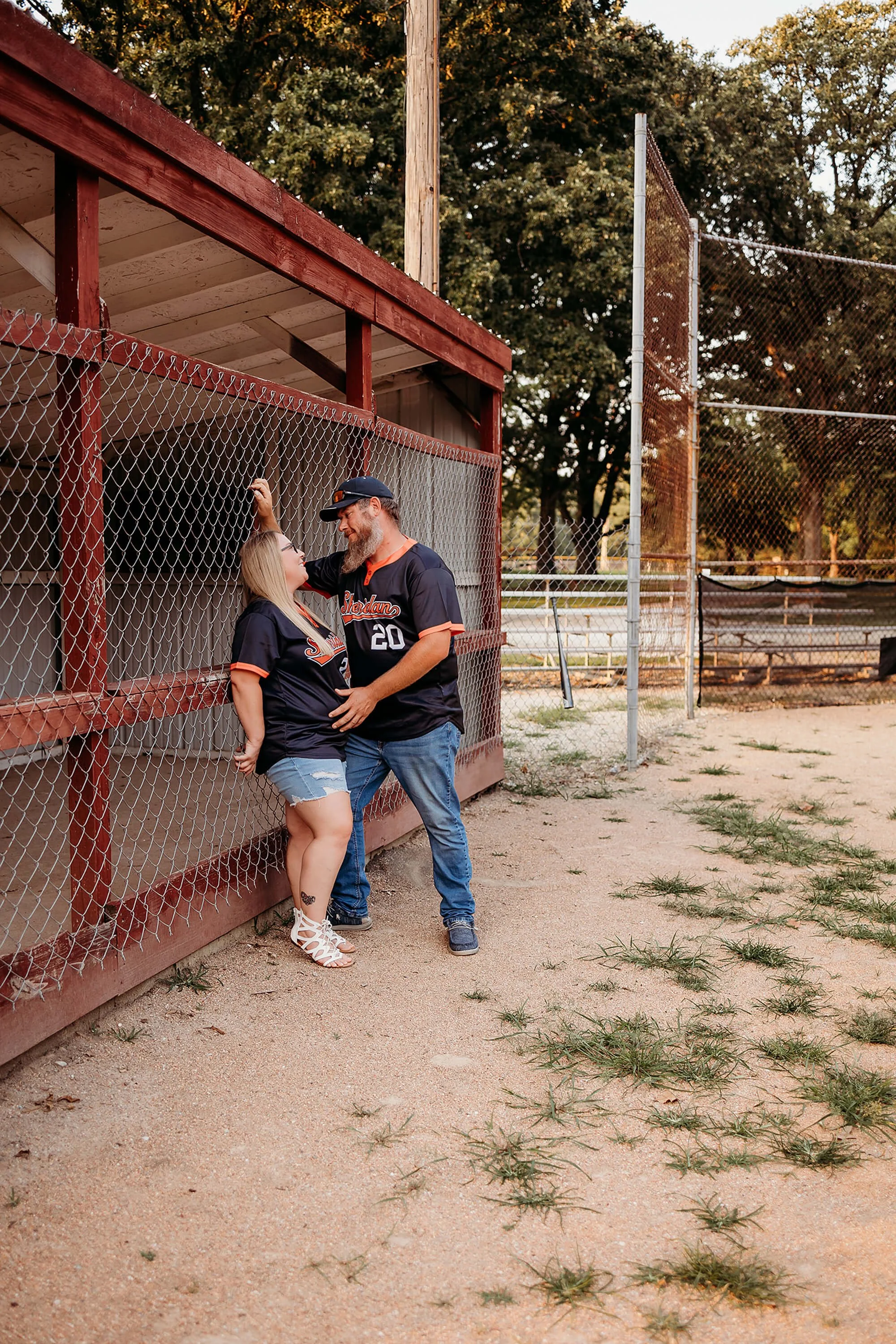 A couple wearing matching baseball jerseys stands at a baseball field, smiling at each other, capturing a warm and intimate pregnancy announcement moment by theheartnarrative Indiana Photographer