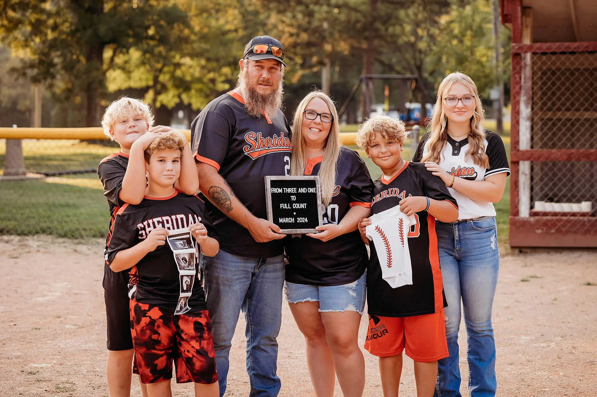 Family in matching baseball jerseys stands together on a field, smiling while holding a pregnancy announcement sign, ultrasound photos, and a baby onesie, captured by theheartnarrative Photographer