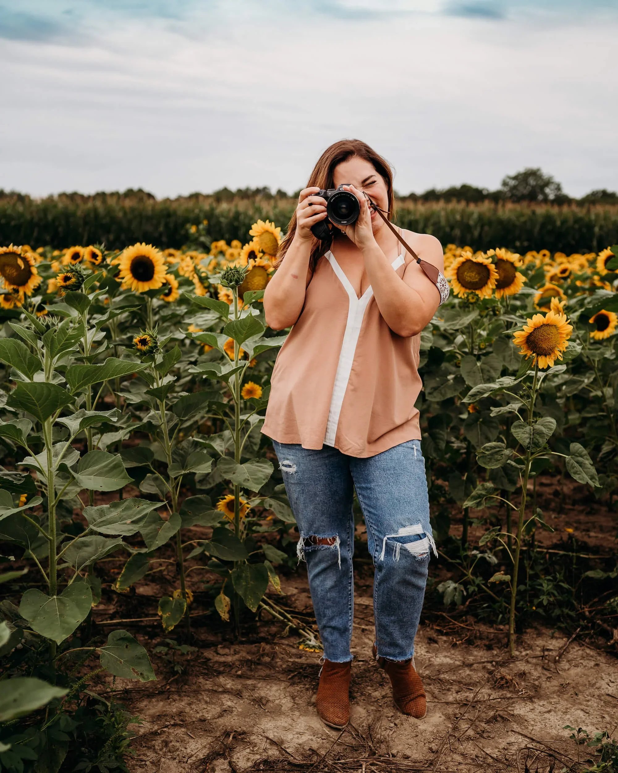 Lifestyle portrait of female photographer smiling with camera in a sunflower field.