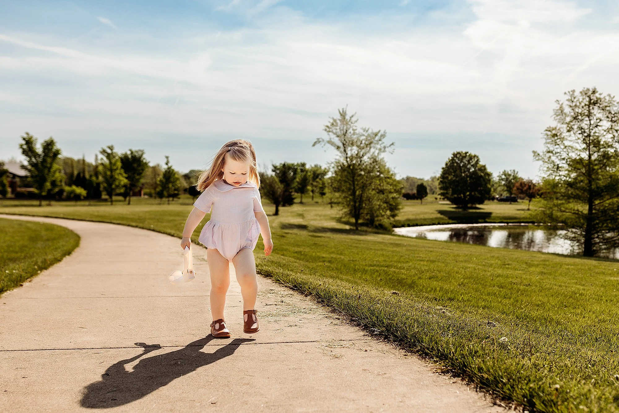 A young girl walking along a paved park pathway holding a small toy, with green grass, trees, and a pond in the background under a partly cloudy sky.