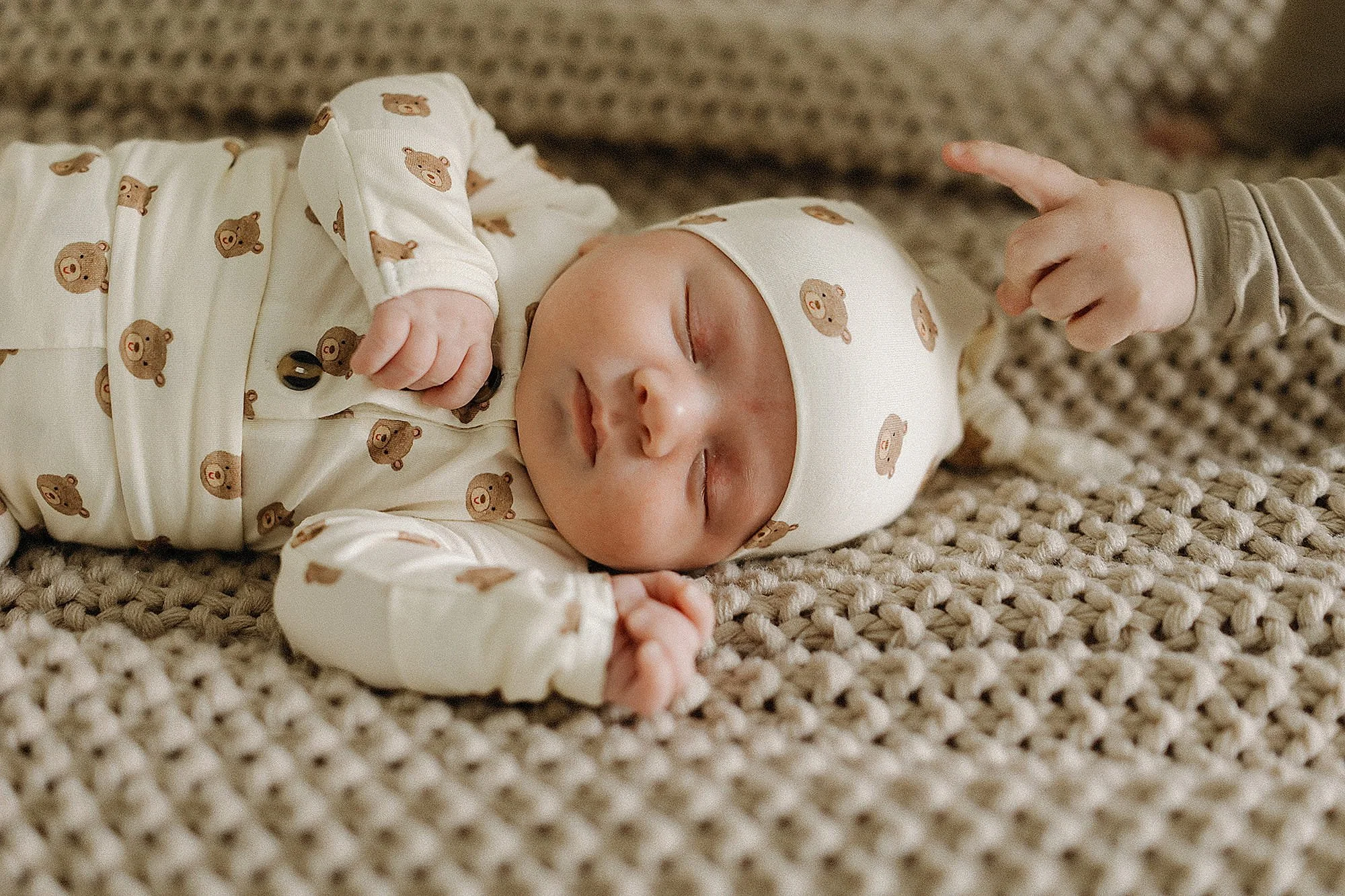 A sleeping baby lying on a textured brown blanket, wearing a cream-colored outfit and hat with brown bear faces, with a small dark object in one hand and a faint smile on the face.