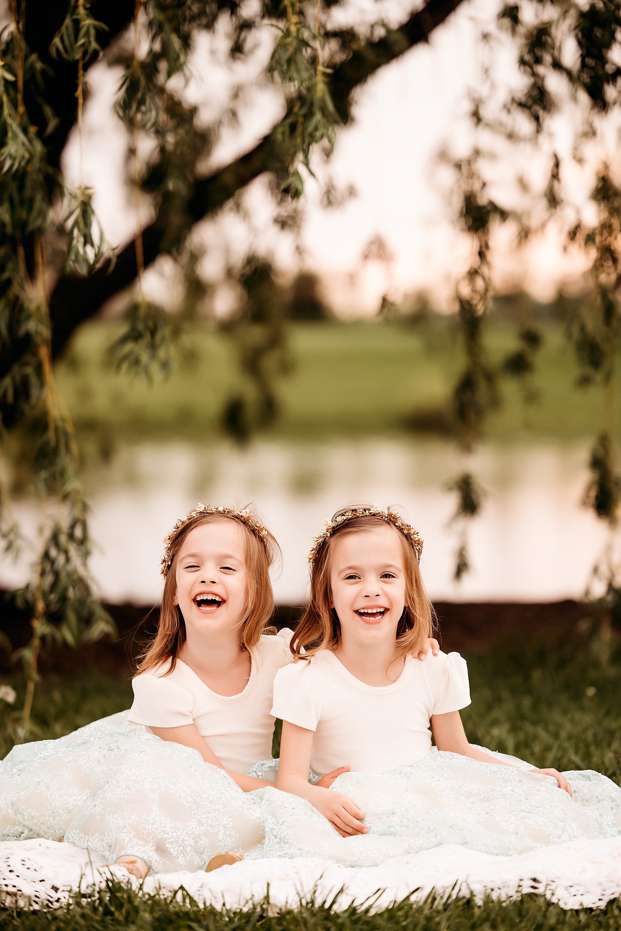 Two little girls in white dresses sitting on the grass during a Mini Session, smiling and enjoying their time together