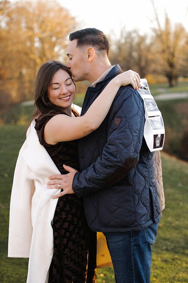 Golden Hour Fall Pregnancy Announcement Photos capturing an expecting couple sharing a tender kiss and embrace, surrounded by glowing autumn foliage and soft evening light.