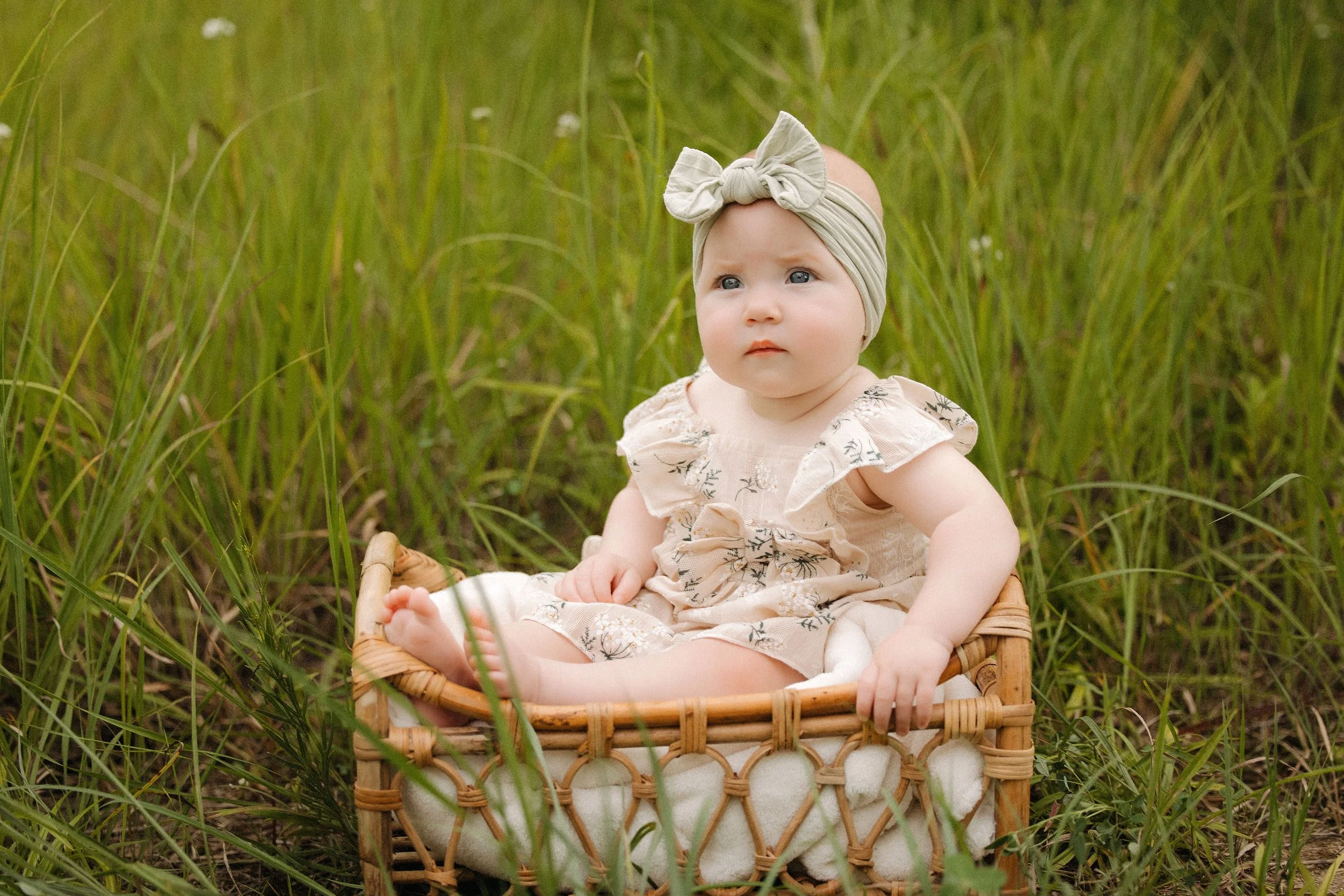 Baby girl sits in a woven basket, smiling softly in a serene outdoor setting during an Extended Family Photo session
