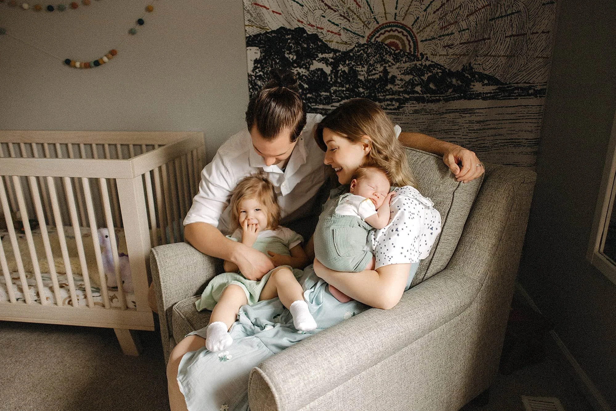 A woman sitting on a grey couch holding an infant, with a young girl sitting beside her. A man is leaning over, engaging with them, in a cozy room with a crib and a large wall hanging with mountain and sun design in the background.