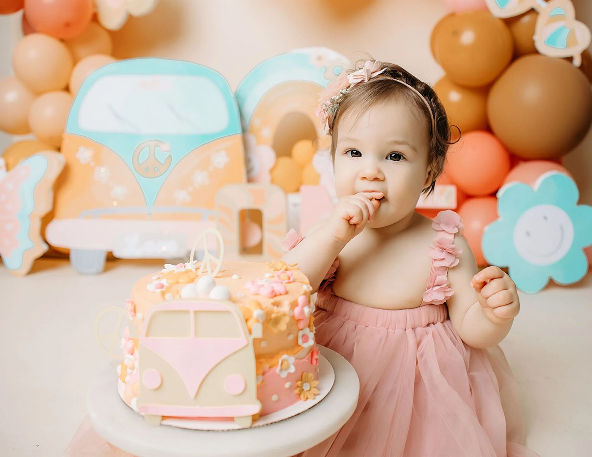 Baby girl in a pink tulle dress and floral headband tasting cake frosting beside a pink and cream birthday cake decorated with a retro VW van and peace sign topper