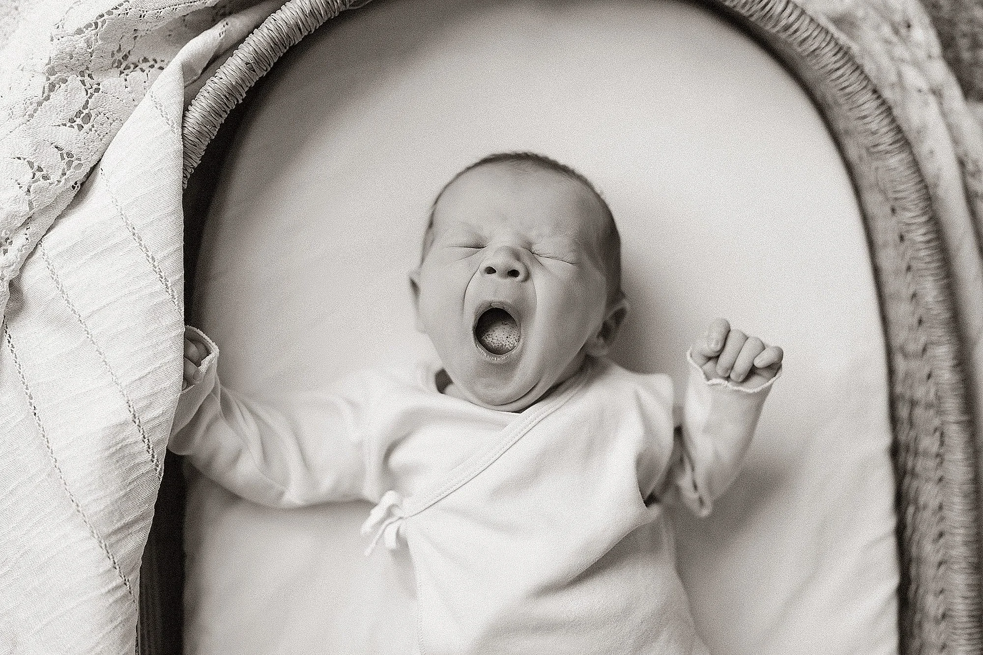 Black and white photo of a yawning newborn baby lying in a bassinet with a lace blanket partially covering the basket.