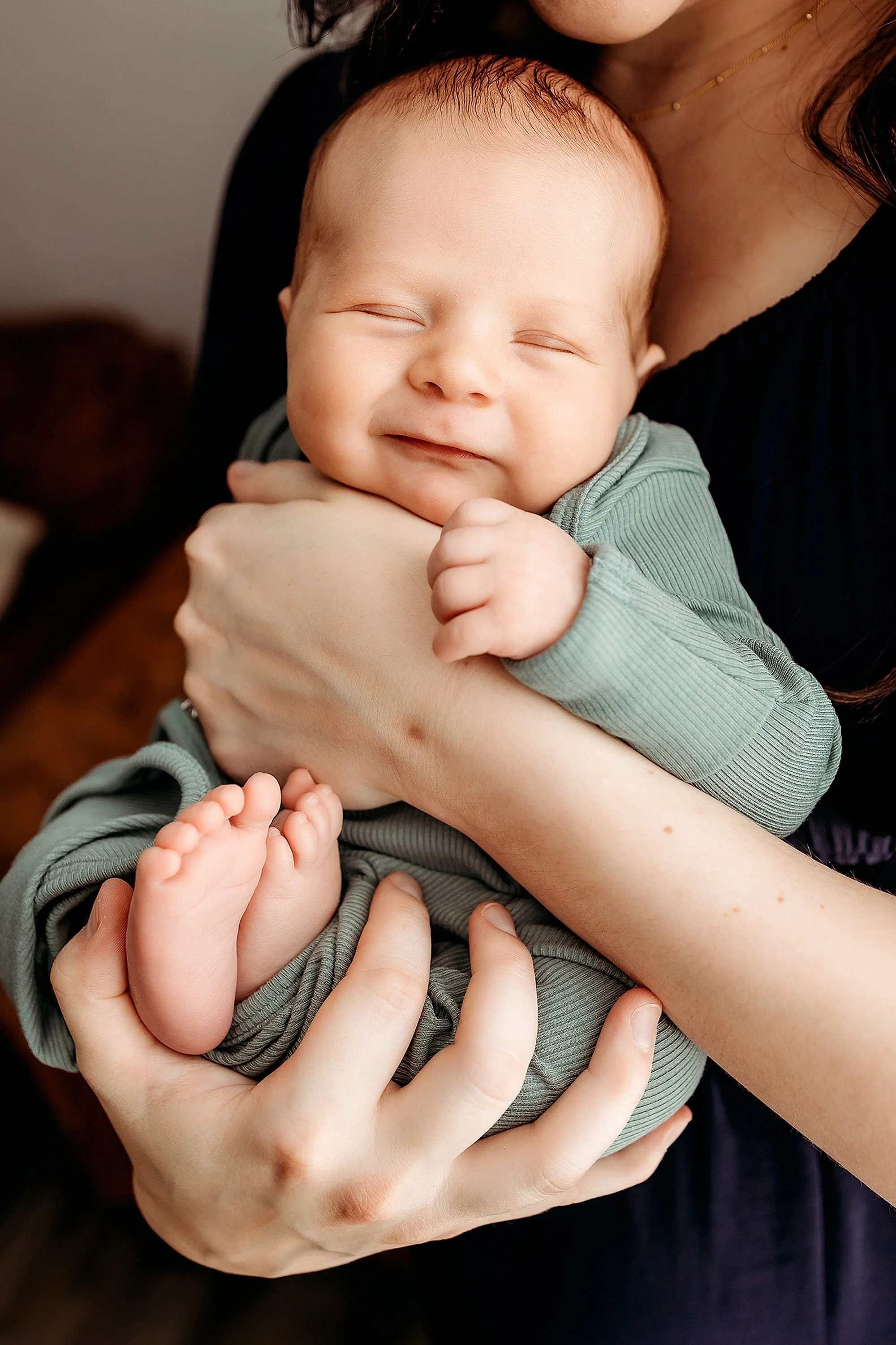 A woman holding a smiling baby in her arms, with the baby resting on her shoulder, against a neutral background.