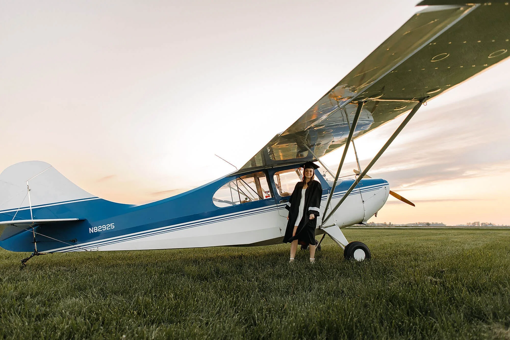 A woman poses next to a small airplane in a field, celebrating her senior photos with a unique aviation backdrop