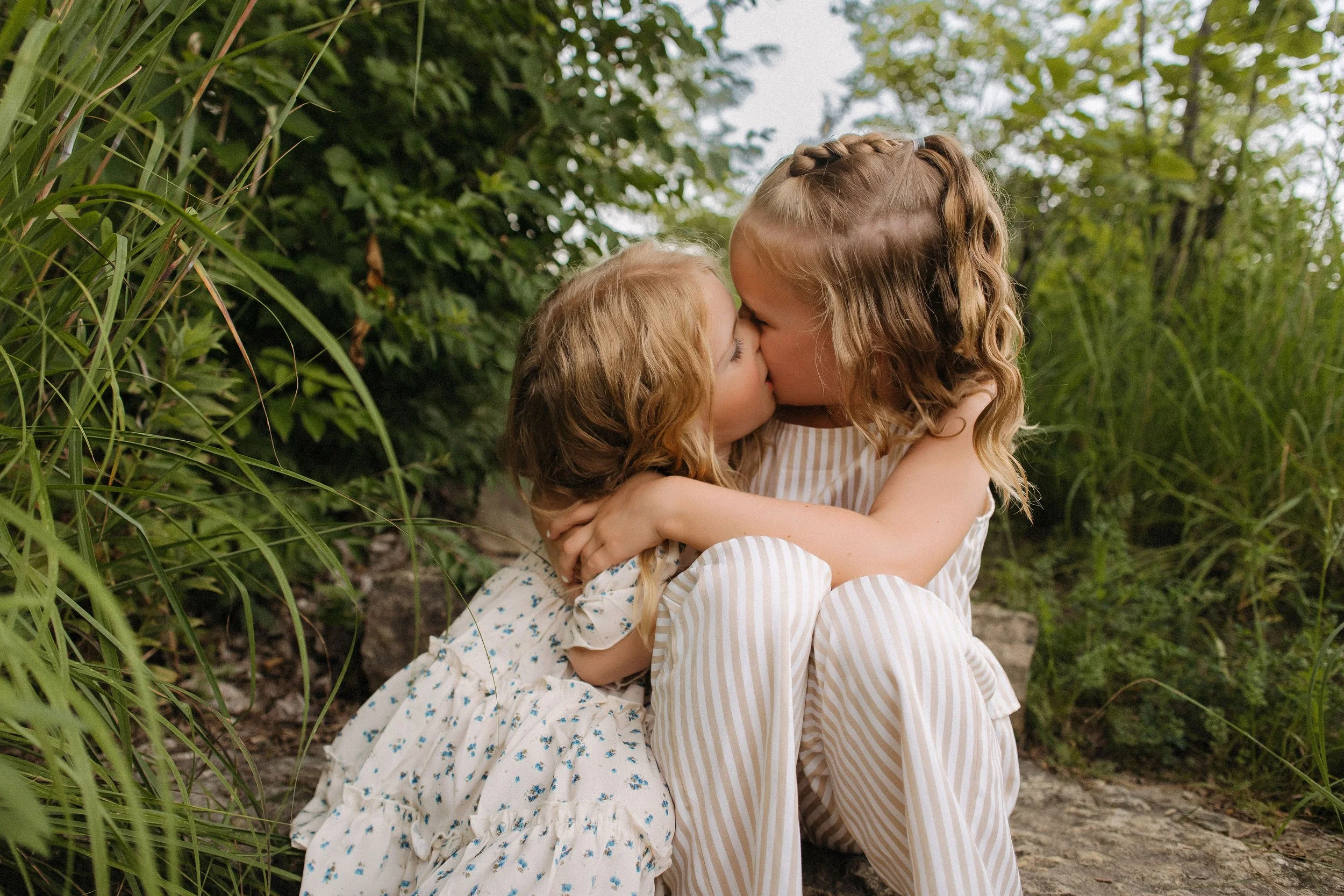 Two little girls share a kiss while sitting on the grass, capturing a sweet moment in an extended family photo session