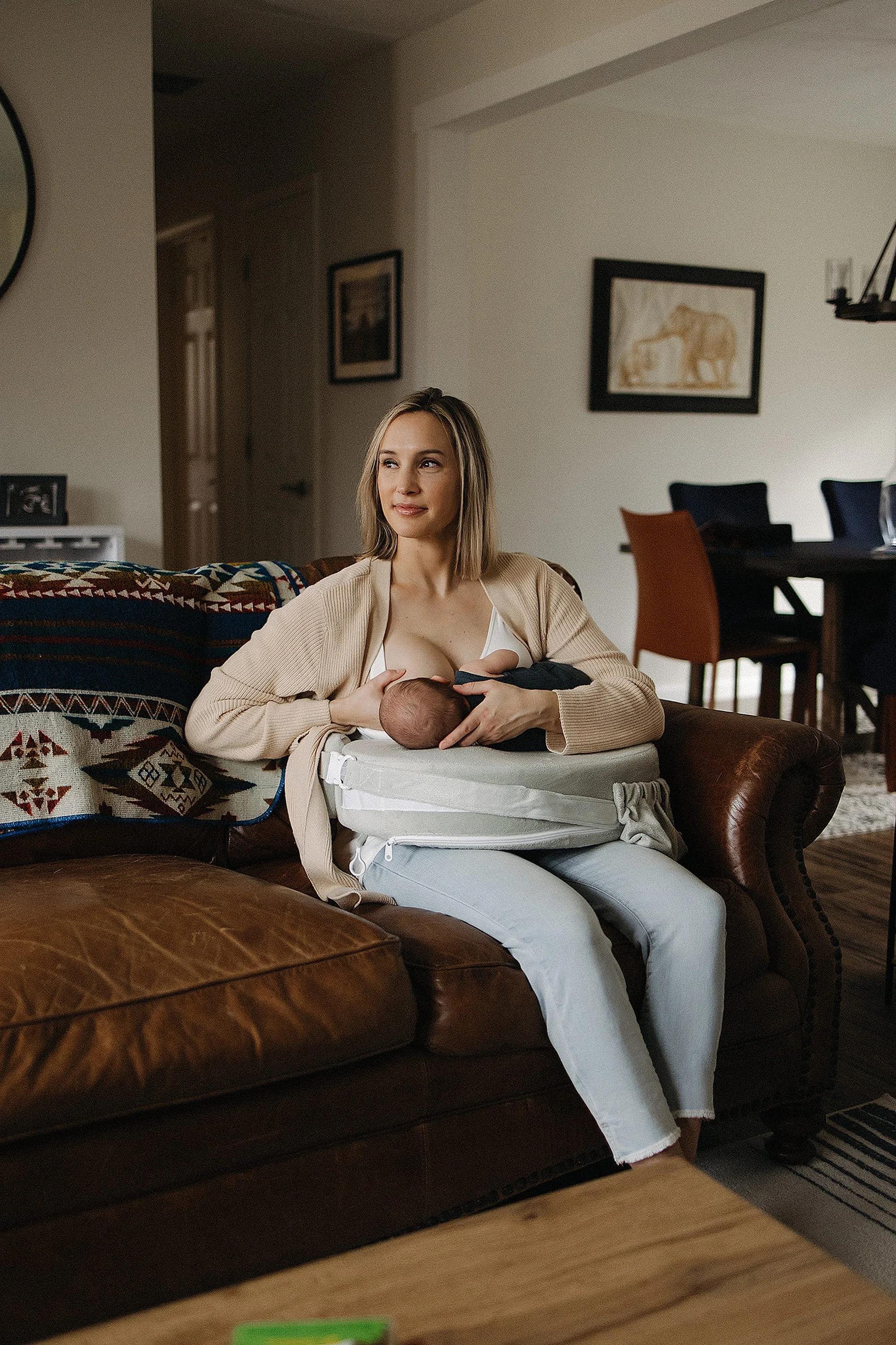 A woman breastfeeding a baby on a brown leather couch in a cozy living room.