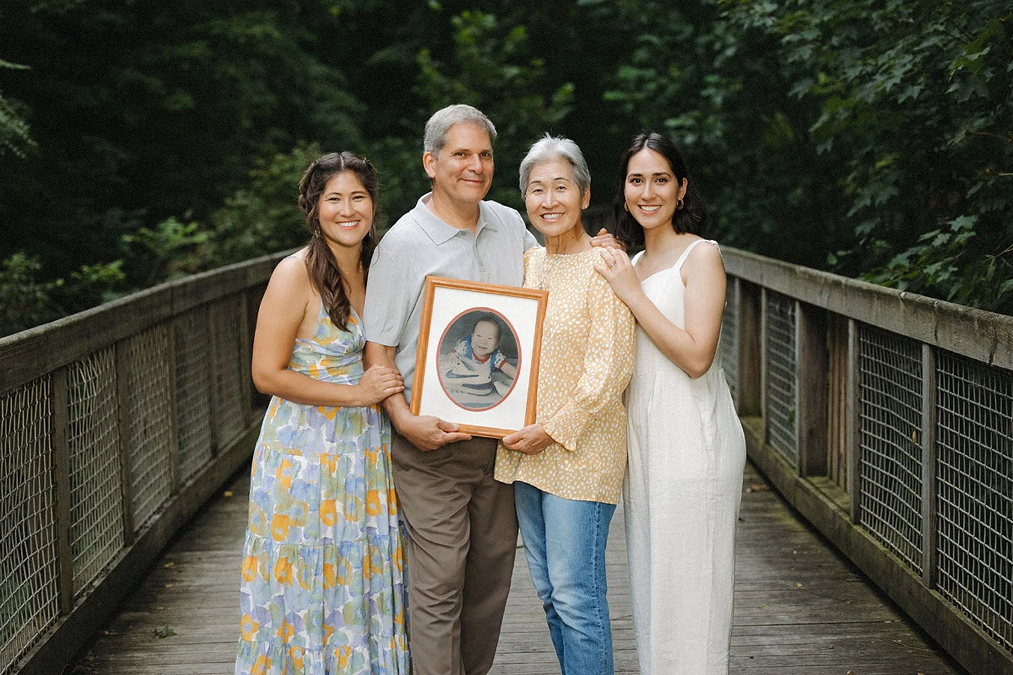 Parents and two adult daughters smiling on a wooden bridge, holding a framed childhood photo, during a Flat Fork Creek Park family photo session