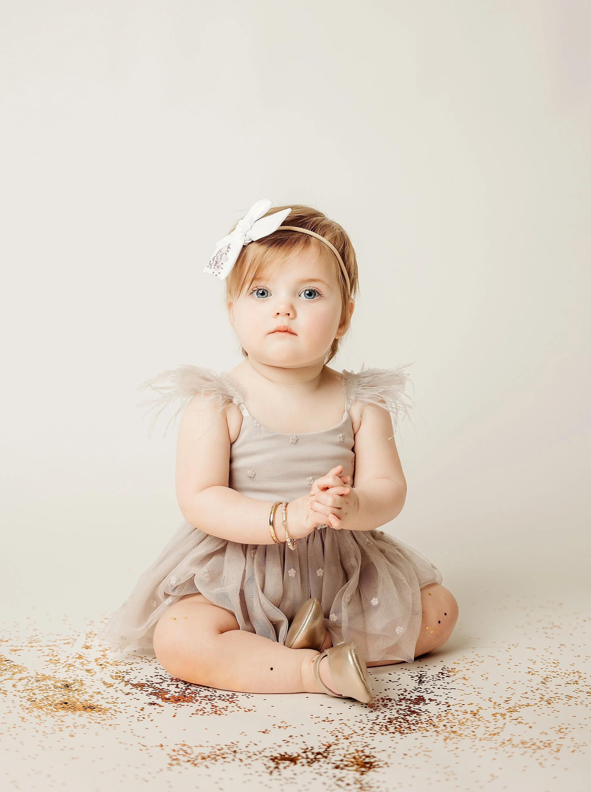 Young girl with blonde hair and blue eyes, wearing a beige tulle dress with feathered shoulders, gold shoes, and a white bow headband, sitting on a white floor sprinkled with gold confetti, looking at the camera.