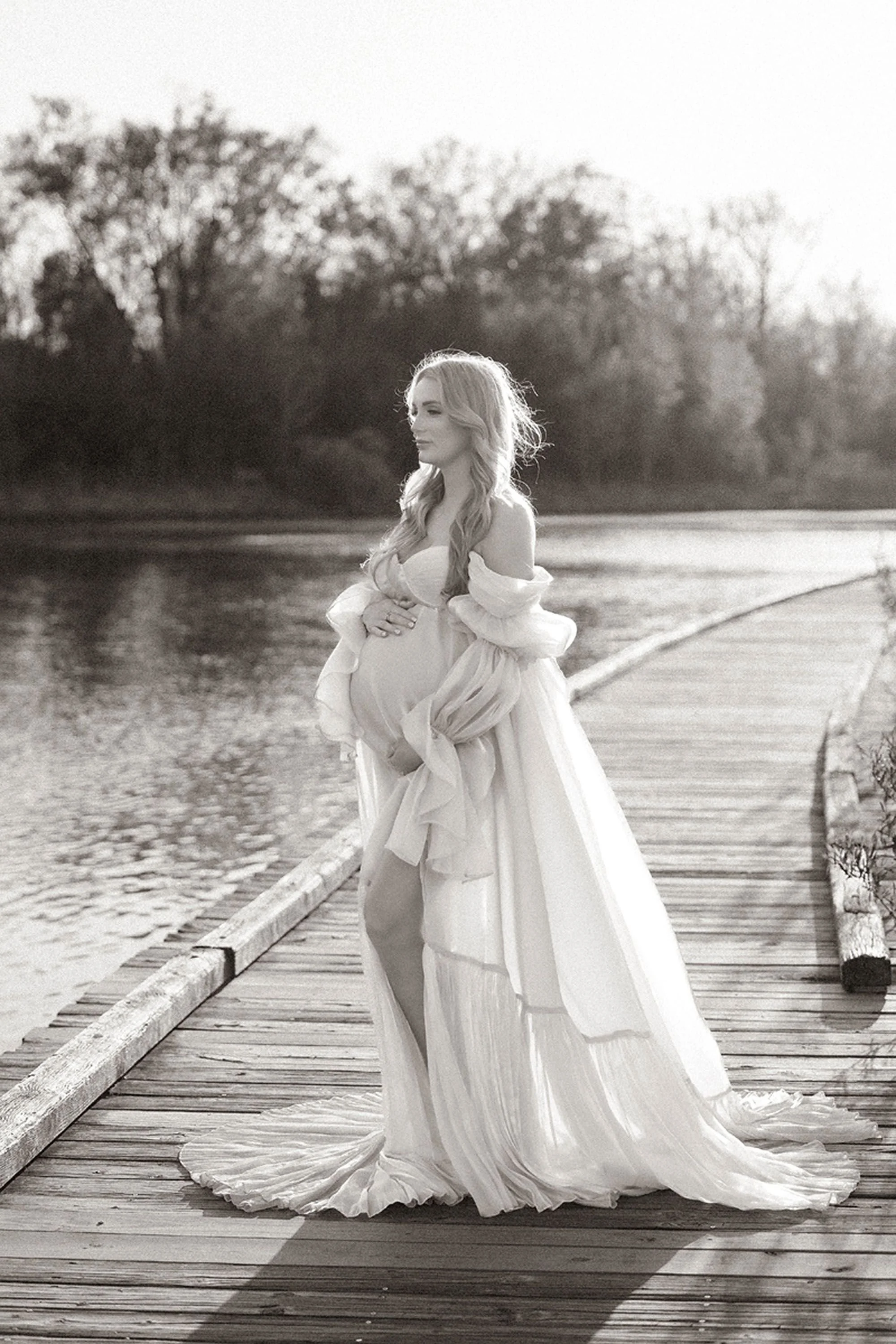 Black and white maternity photos in Central Indiana featuring a pregnant woman in profile on a wooden boardwalk beside a still lake, gown flowing behind her