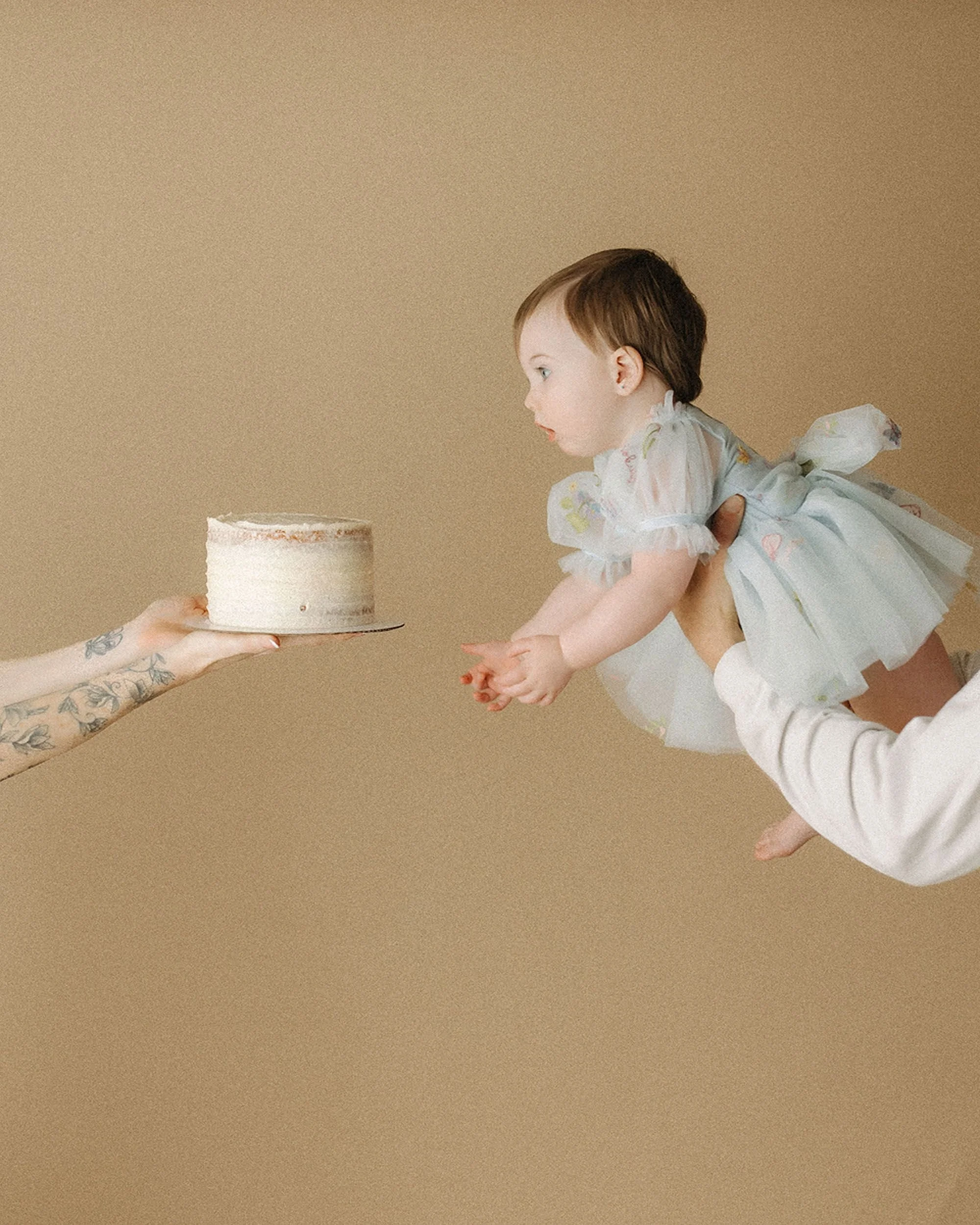 Simple Cake Smash Photos of a baby girl in a white dress reaching toward a small birthday cake, creating a playful and natural first-birthday moment.