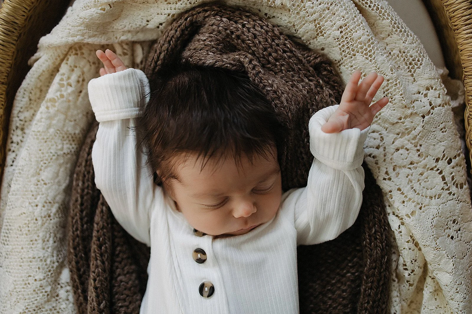 A sleeping baby with dark hair, wearing a white ribbed onesie with black buttons, lying on a cream crochet blanket, with a brown knitted blanket partially covering them.