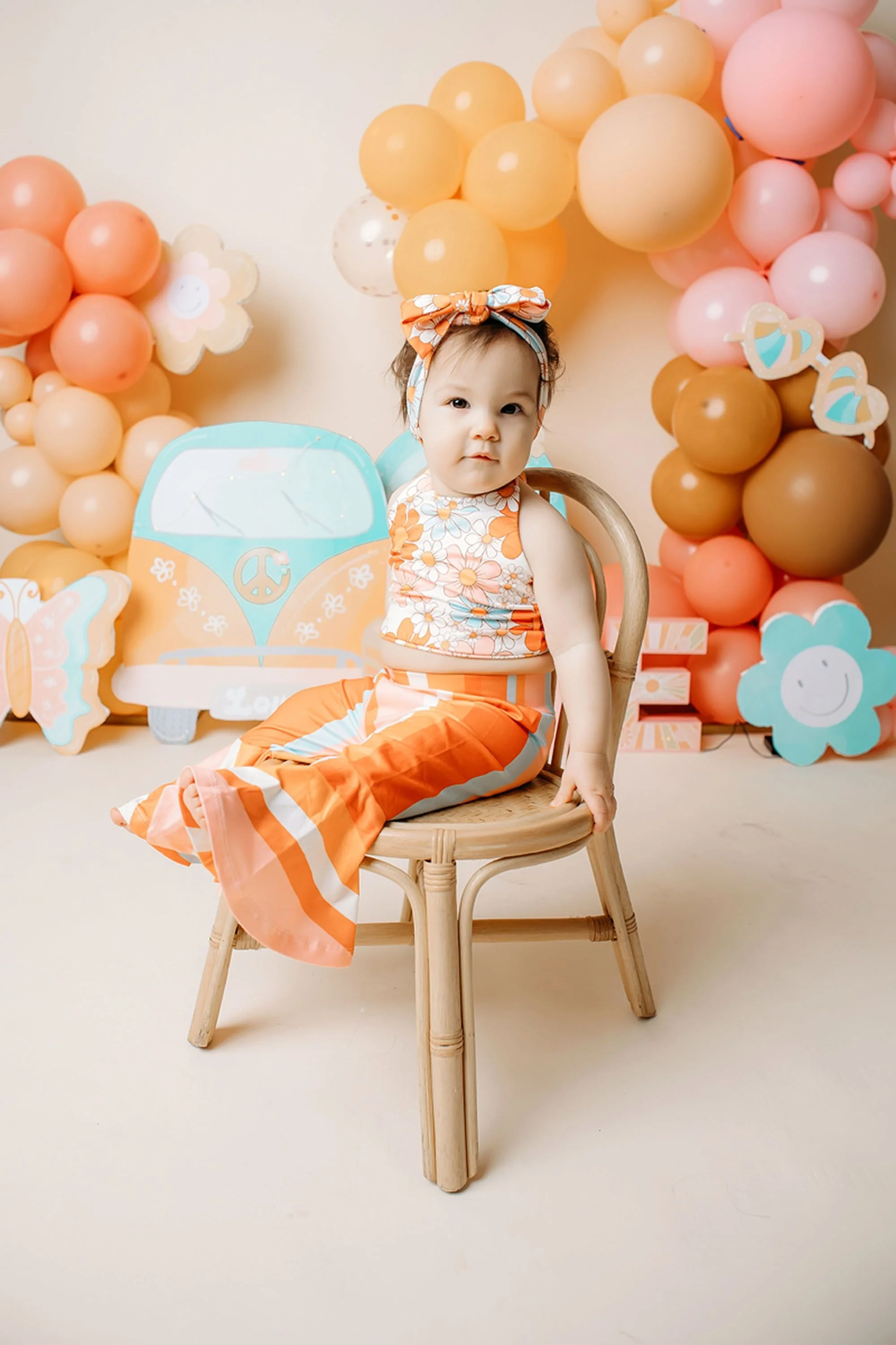 A baby girl poses on a wooden chair in a cheerful Groovy One cake smash backdrop featuring pastel balloons, rainbow accents, flowers, and a colorful retro outfit.