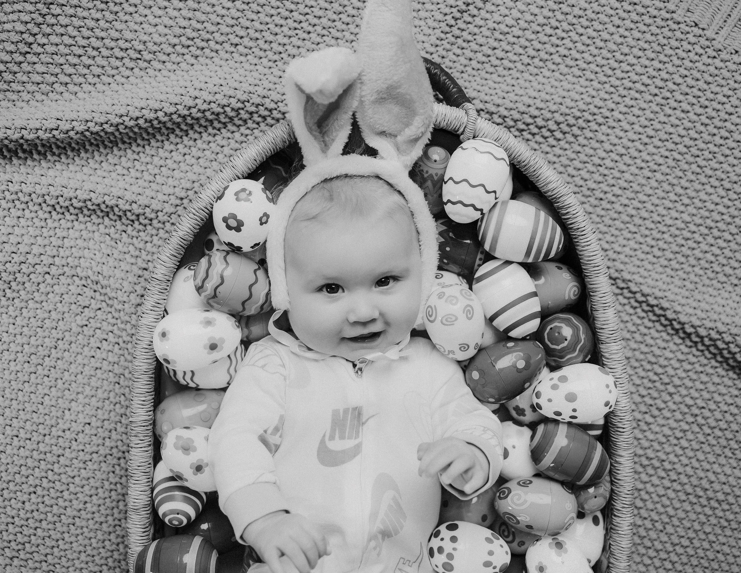 baby in a basket surrounded by toys eggs, wearing a cute bunny hat for a 6-month milestone photoshoot.