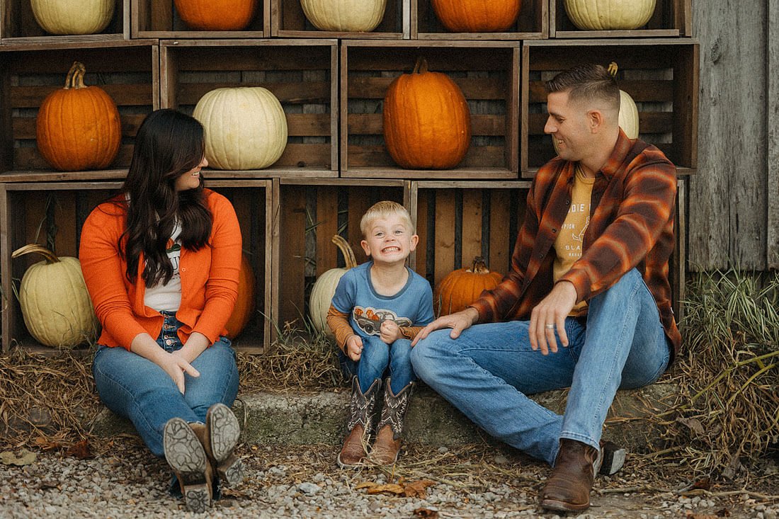 Family of three sits together beside wooden crates displaying orange and white pumpkins during their fall visit to Dull’s Pumpkin Harvest