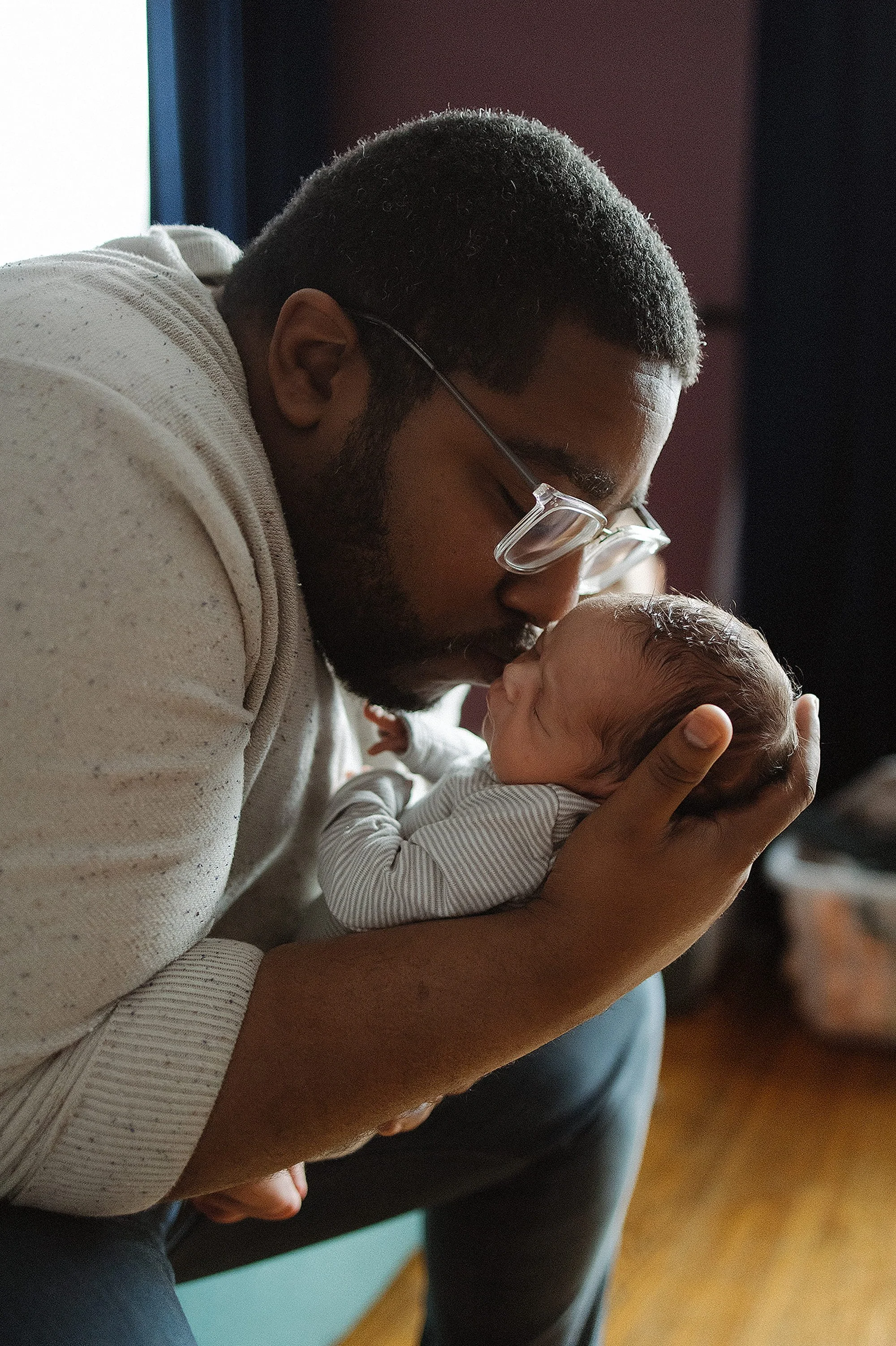 A father with glasses holding a newborn baby close to his face, touching foreheads, near a window in a warmly lit room during an in home lifestyle photoshoot with a newborn.