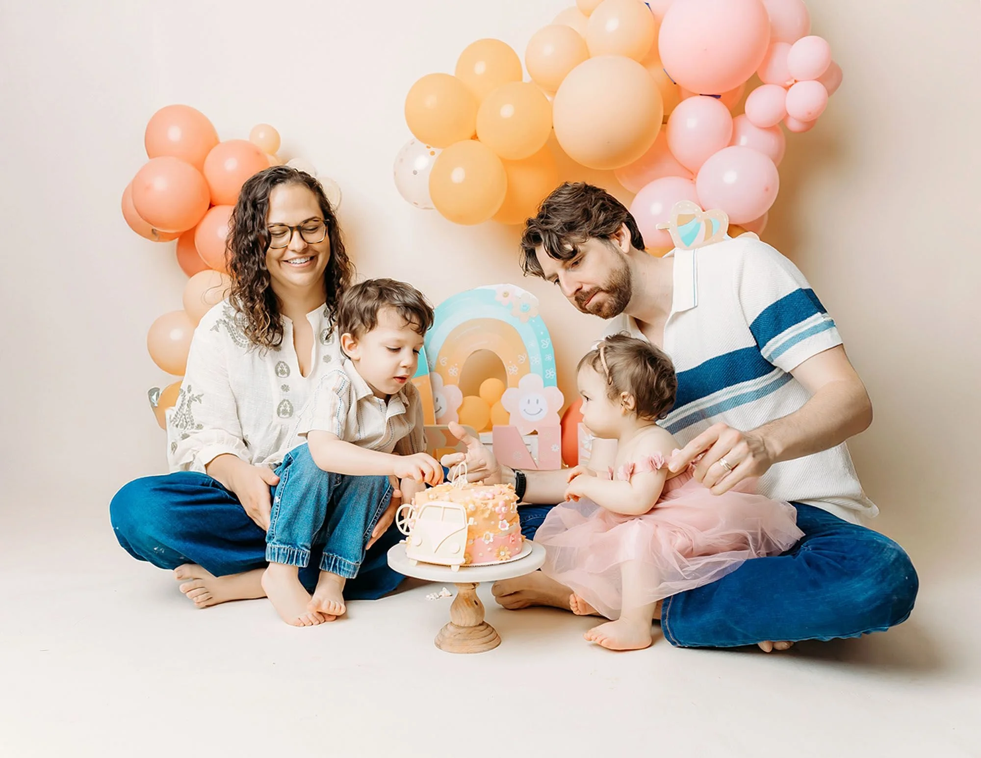 Happy family sitting together on a cream backdrop with pink, peach, and yellow balloon garlands, gathered around a groovy-themed first birthday cake with a retro van topper for a cake smash photo session