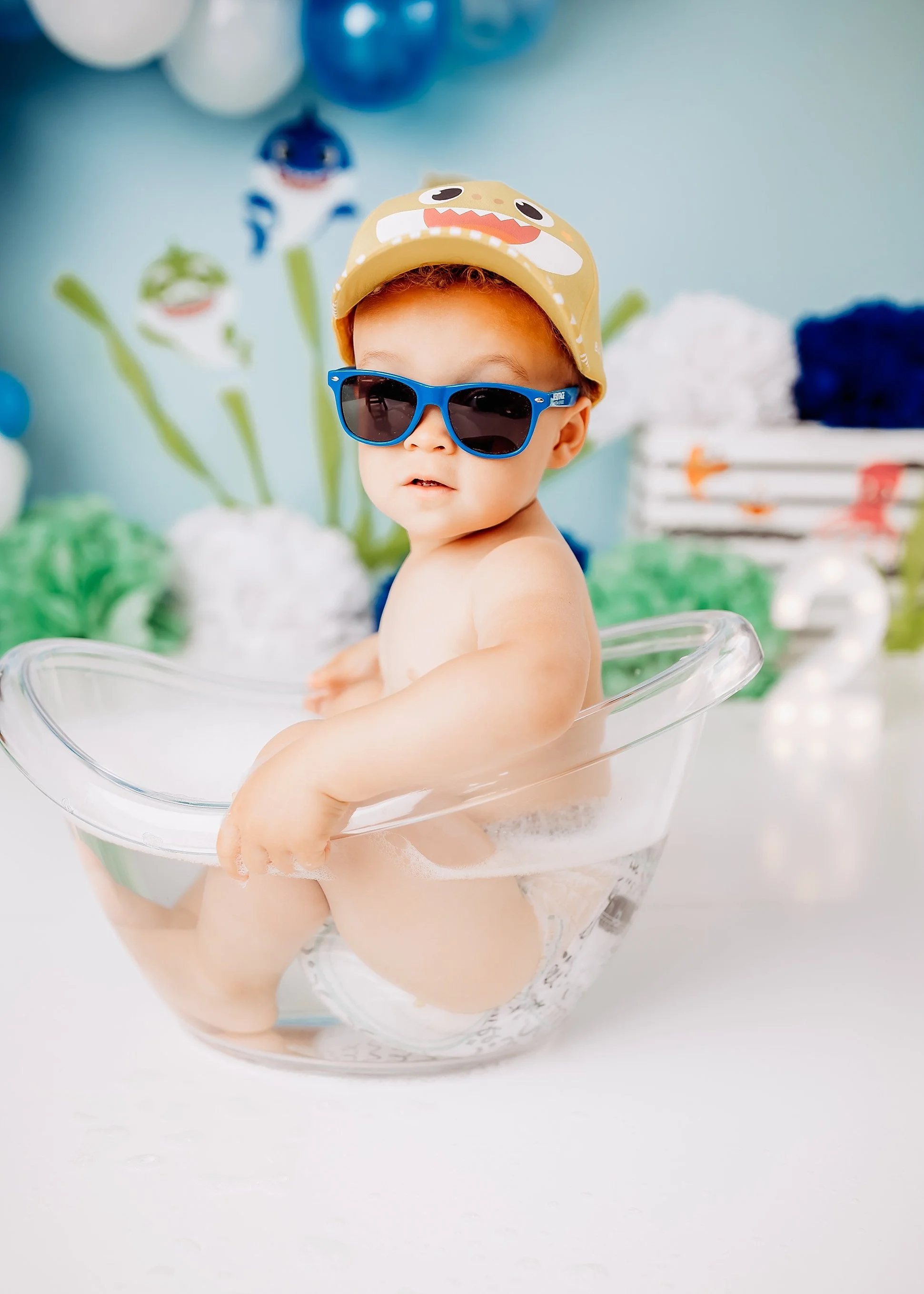 A baby wearing sunglasses and a hat, sitting in a clear plastic bathtub with soap bubbles, in a decorated room with blue, white, and green balloons and wall decorations.