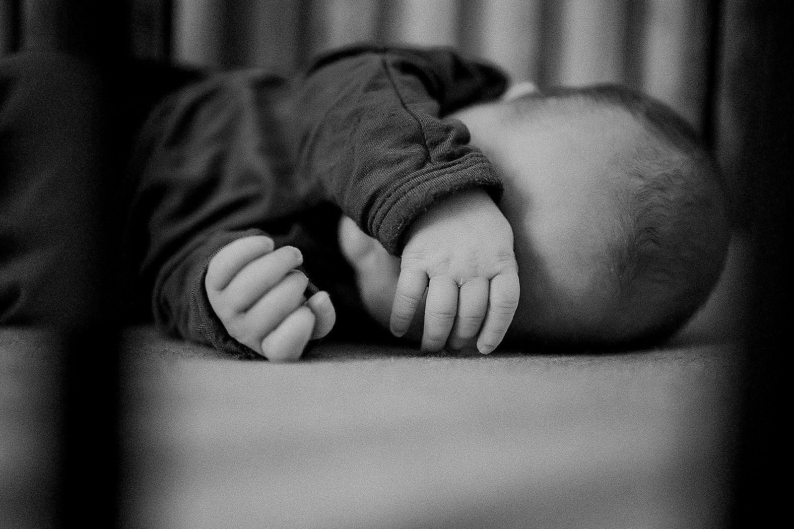 A baby laying on a surface, viewed from the side in black and white, with its hands grasped together near its face.