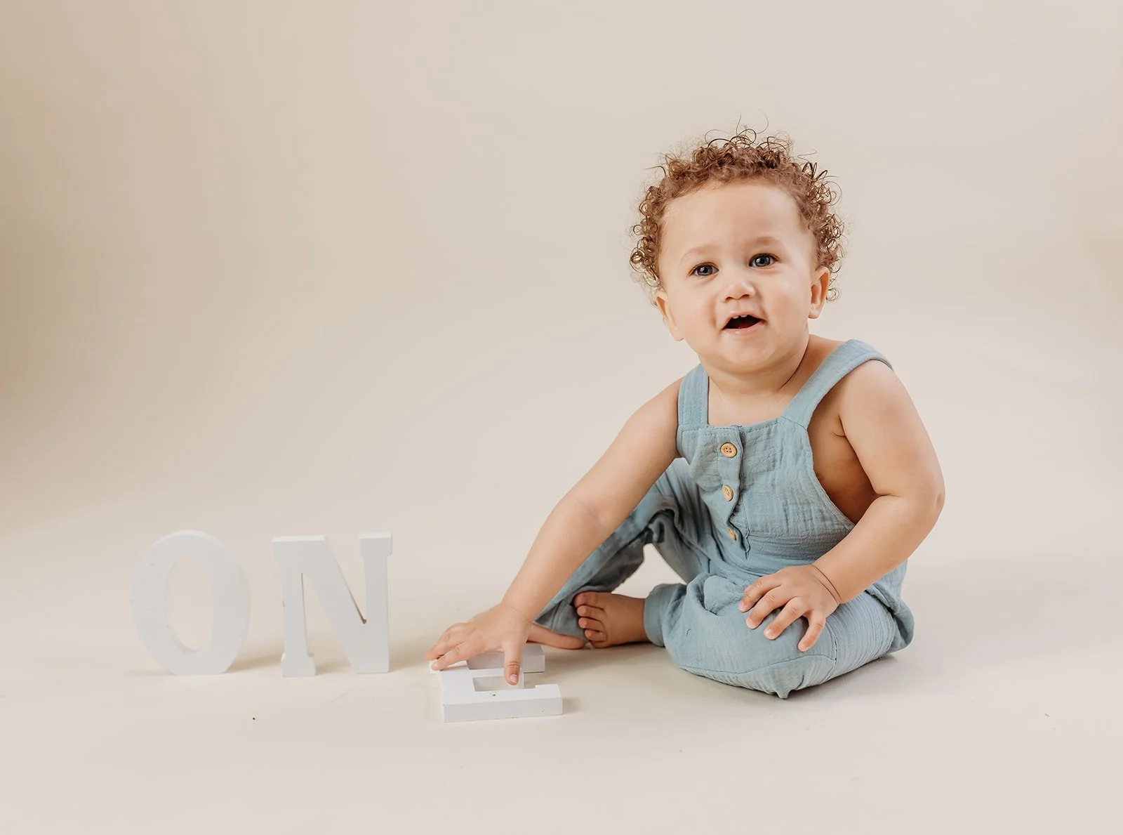 A young toddler with curly hair, wearing a light blue sleeveless outfit, sitting on a light-colored surface. The child is reaching towards a white decorative letter 'E' with their left hand. Two other white letters, 'O' and 'N', are positioned to the