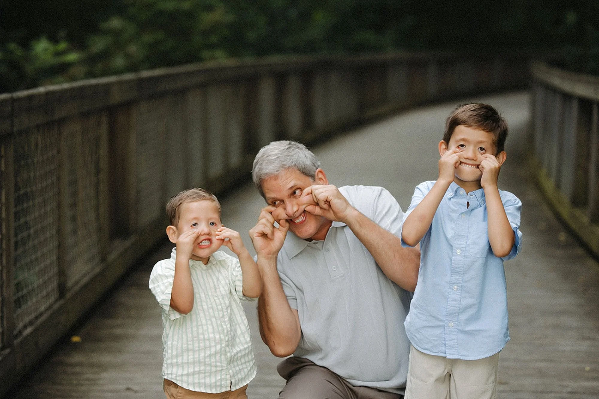 Grandfather making silly faces with his two grandsons on a wooden boardwalk during a Flat Fork Creek Park family photo session, playful candid moment
