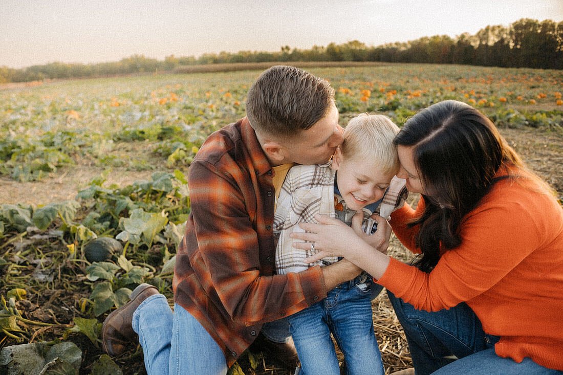 Parents cuddle and kiss their laughing young son in the pumpkin patch while enjoying a playful family moment at Dull’s Pumpkin Harvest