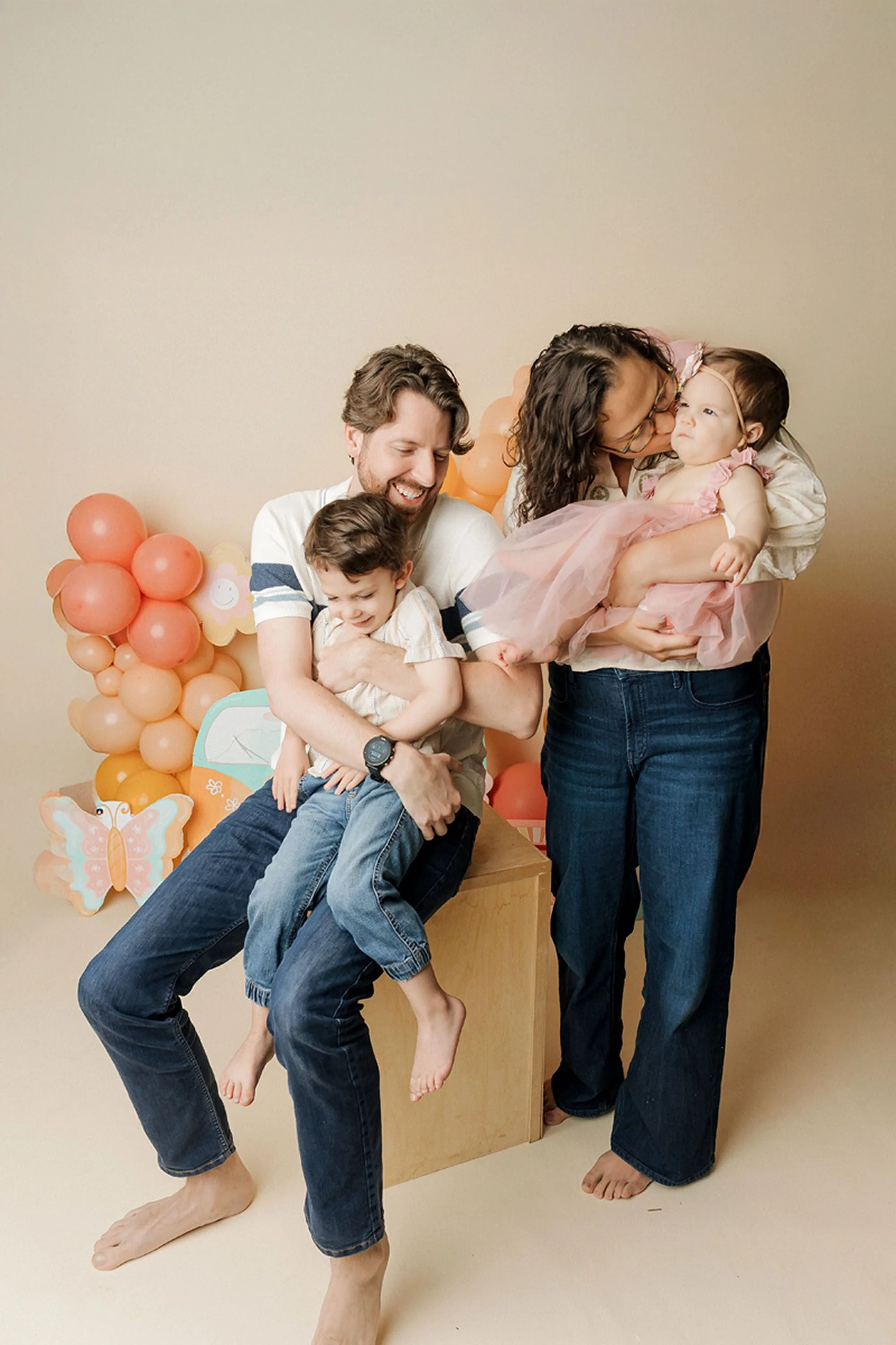 Parents holding their two children on a cream backdrop with pink and peach balloon garlands — dad hugging his laughing toddler son, mom kissing her baby daughter in a pink tulle outfit