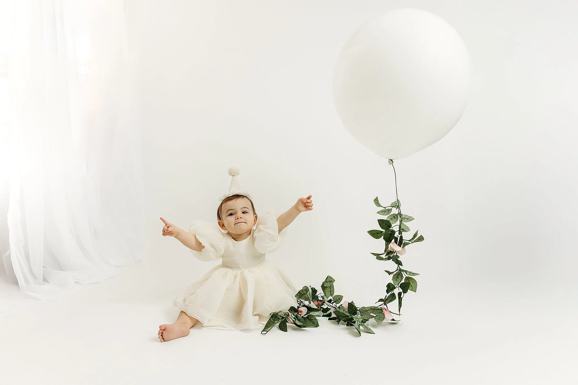A young girl dressed in a white dress and wearing a party hat sitting on the floor next to a large white balloon with green leaves and pink flowers attached to its string, set against a white background.
