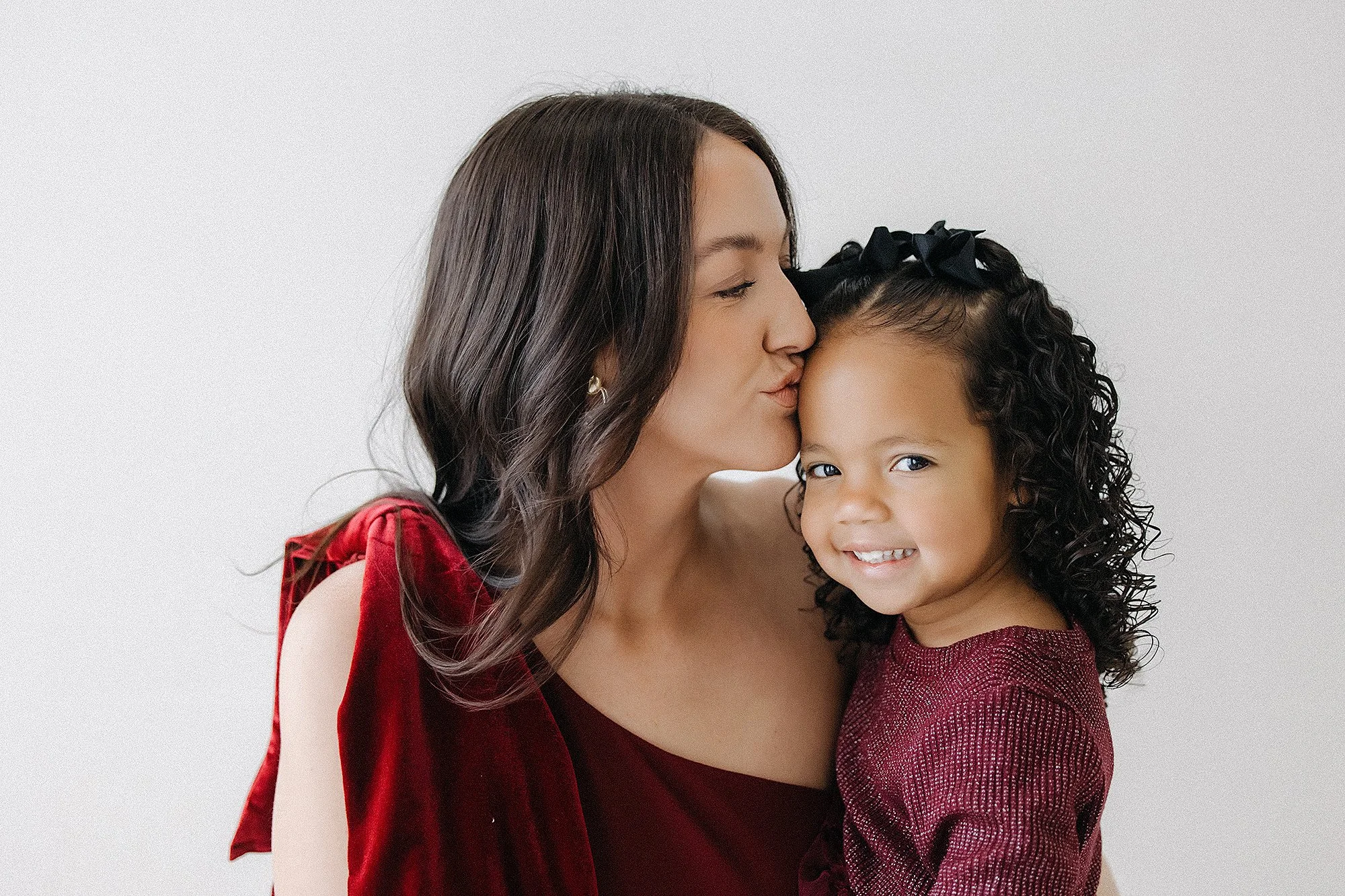 Mother kissing her smiling daughter during a cozy indoor portrait, wearing elegant burgundy outfits that inspire what to wear for family photos with timeless colors, warmth, and heartfelt connection