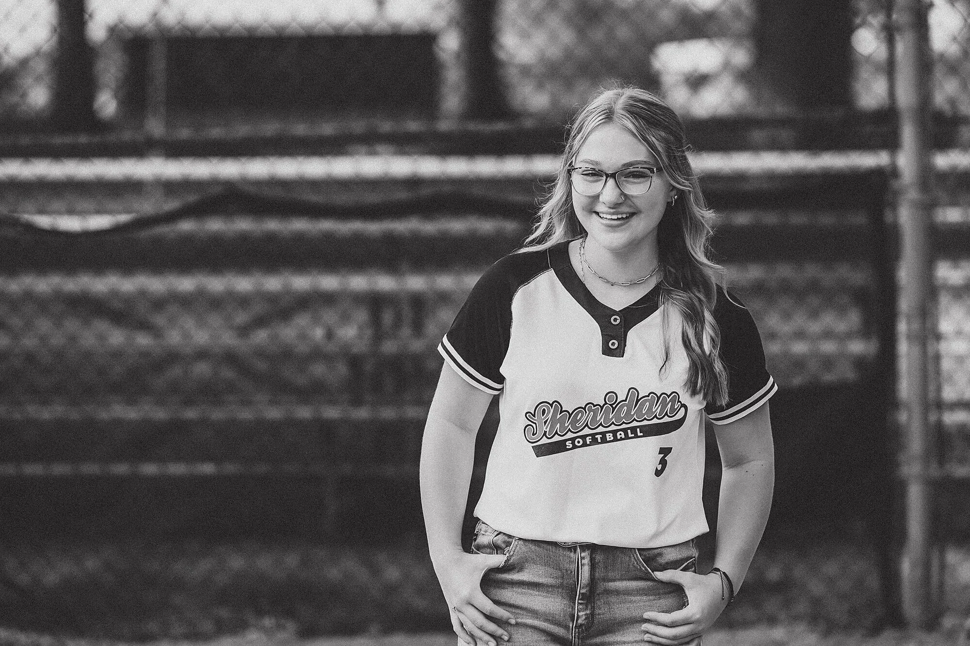 A smiling teenage girl wearing glasses stands outdoors, dressed in a “Sheridan Softball” jersey and jeans, in a black-and-white portrait.