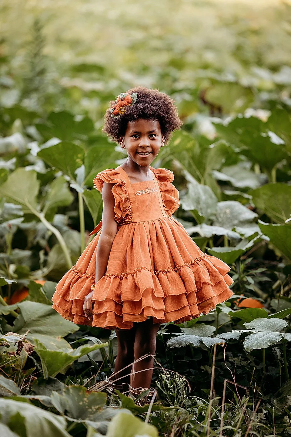 girl in orange boutique dress stands in the middle of a pumpkin patch at dulls tree farm, smiling a the camera
