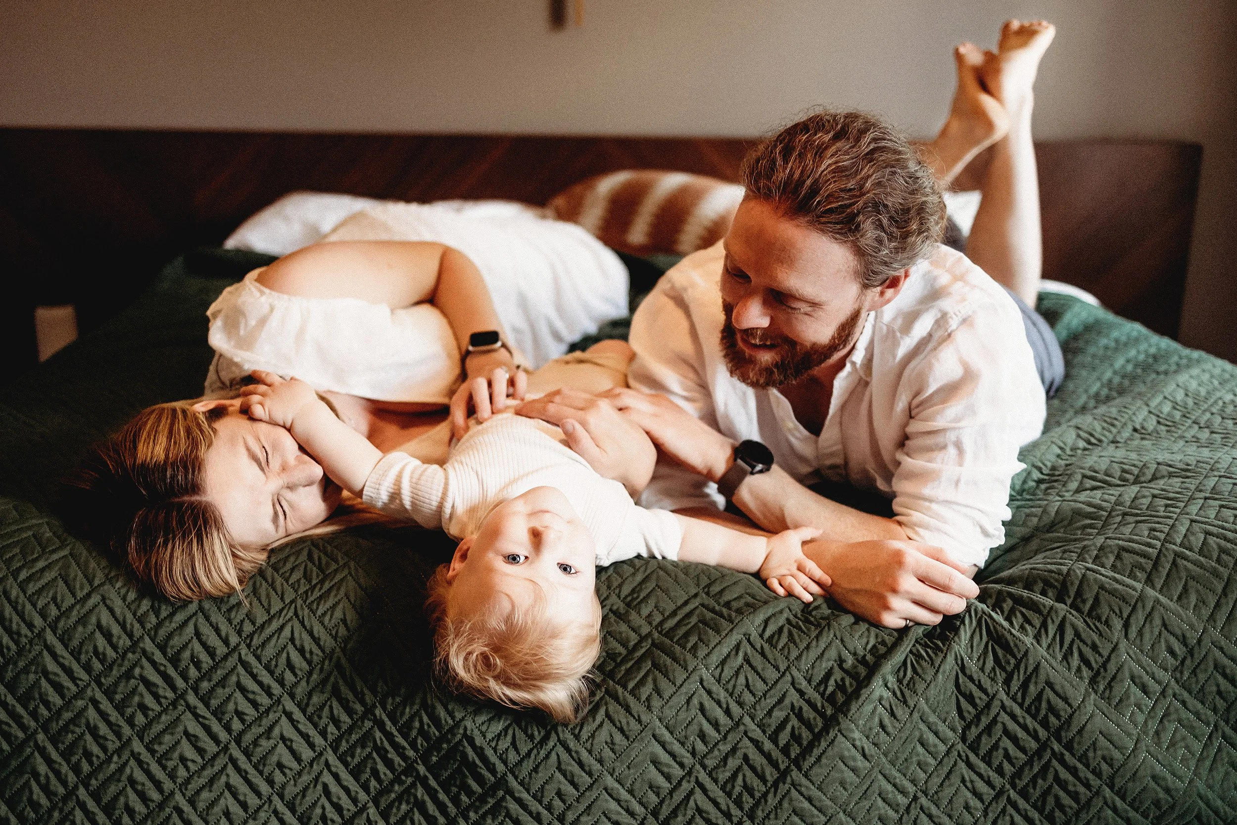 A family of four having fun on a bed, with a woman lying down, a toddler in a white shirt lying on top, and a man playing with the child, all smiling and laughing.