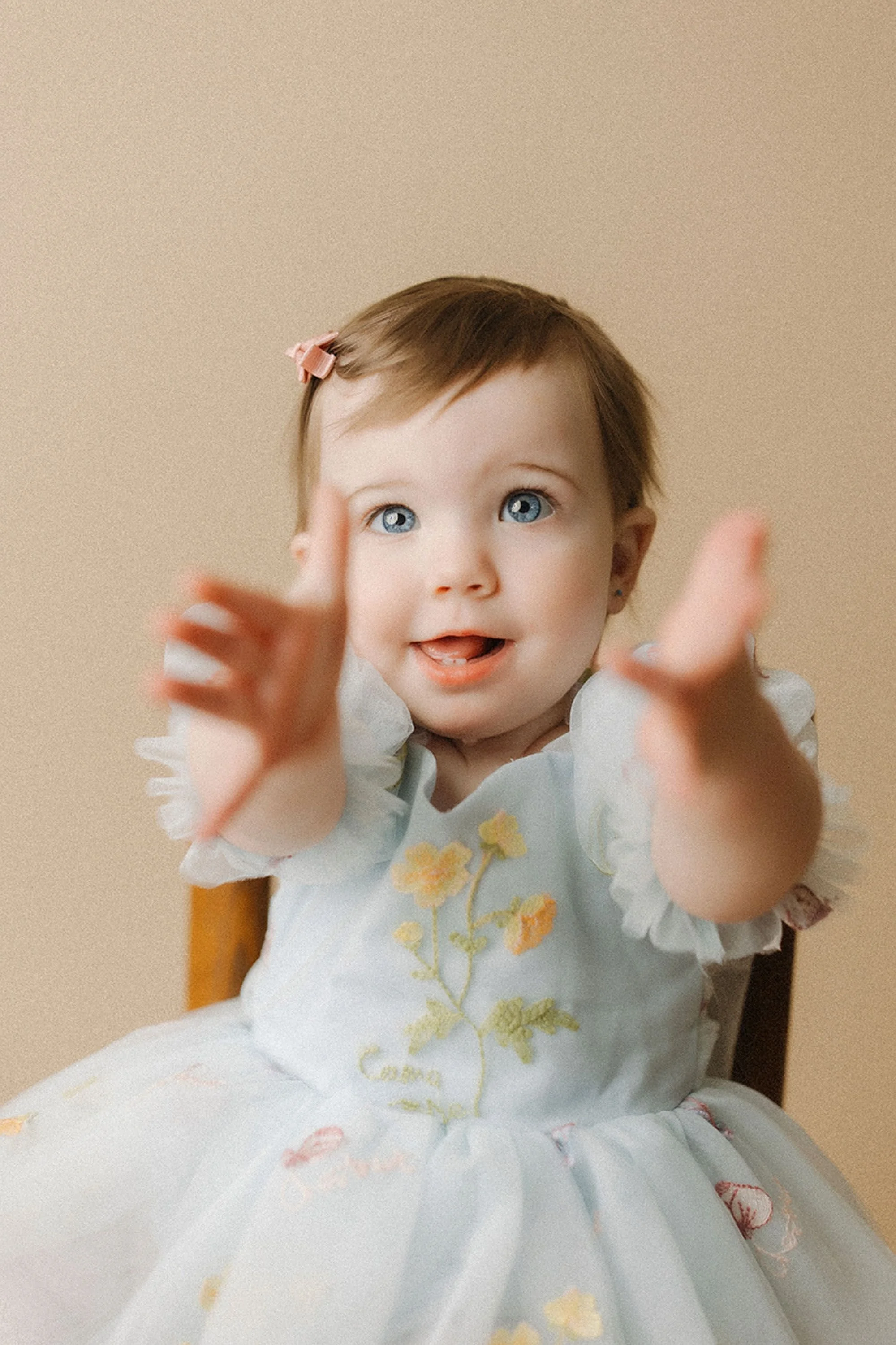 Simple Cake Smash Photos featuring an adorable baby girl with bright blue eyes stretching out her hands while wearing a soft blue floral dress, creating a timeless first birthday portrait.
