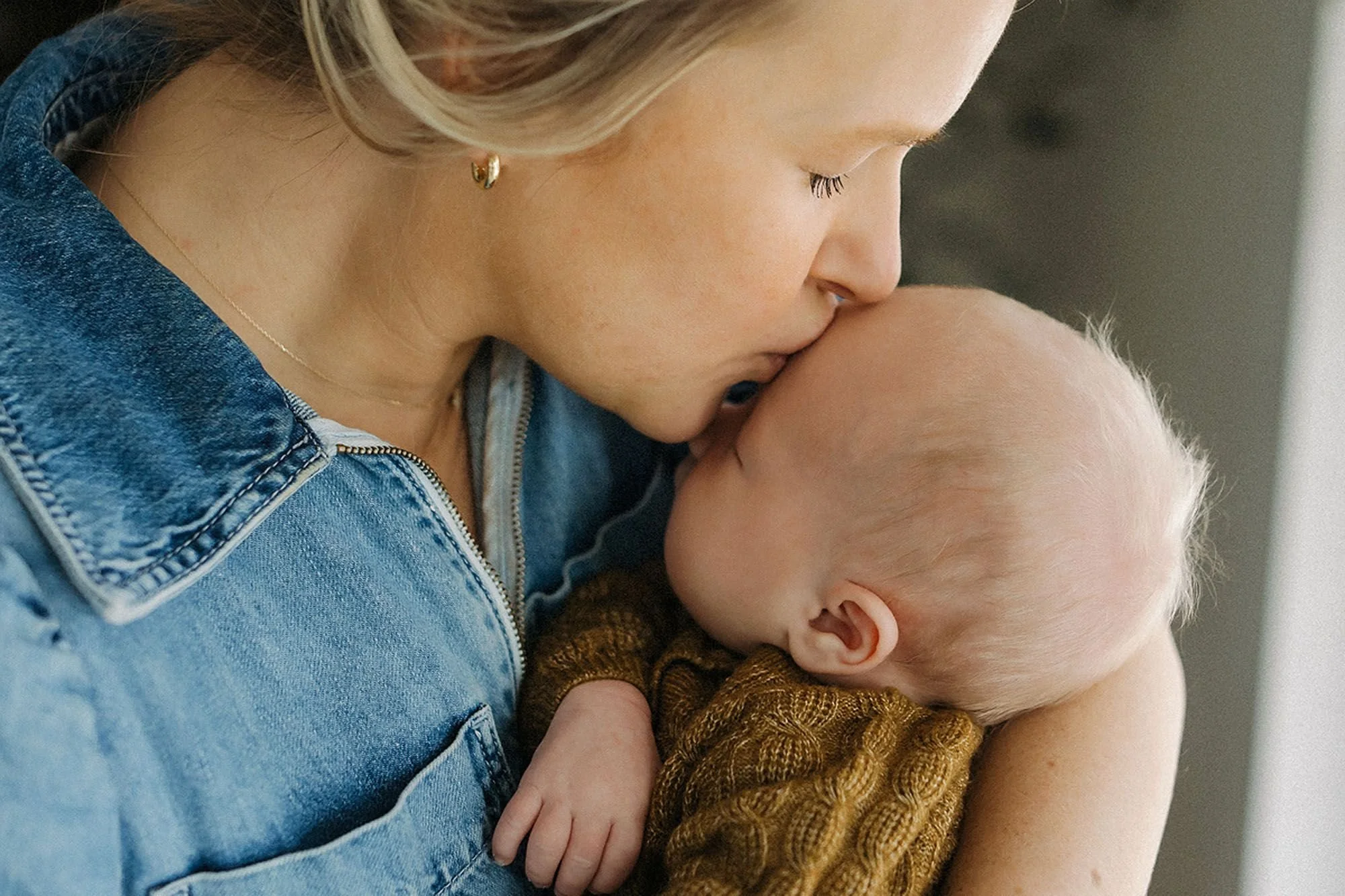 Tender lifestyle newborn photography of a loving mother gently kissing her baby's forehead, capturing an intimate bonding moment, and timeless in-home newborn family portraits