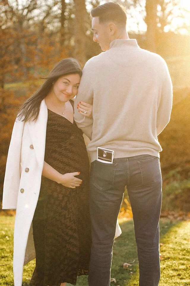 Expecting couple shares a joyful moment outdoors, with an ultrasound photo and gentle embrace during a warm golden-hour pregnancy announcement session.