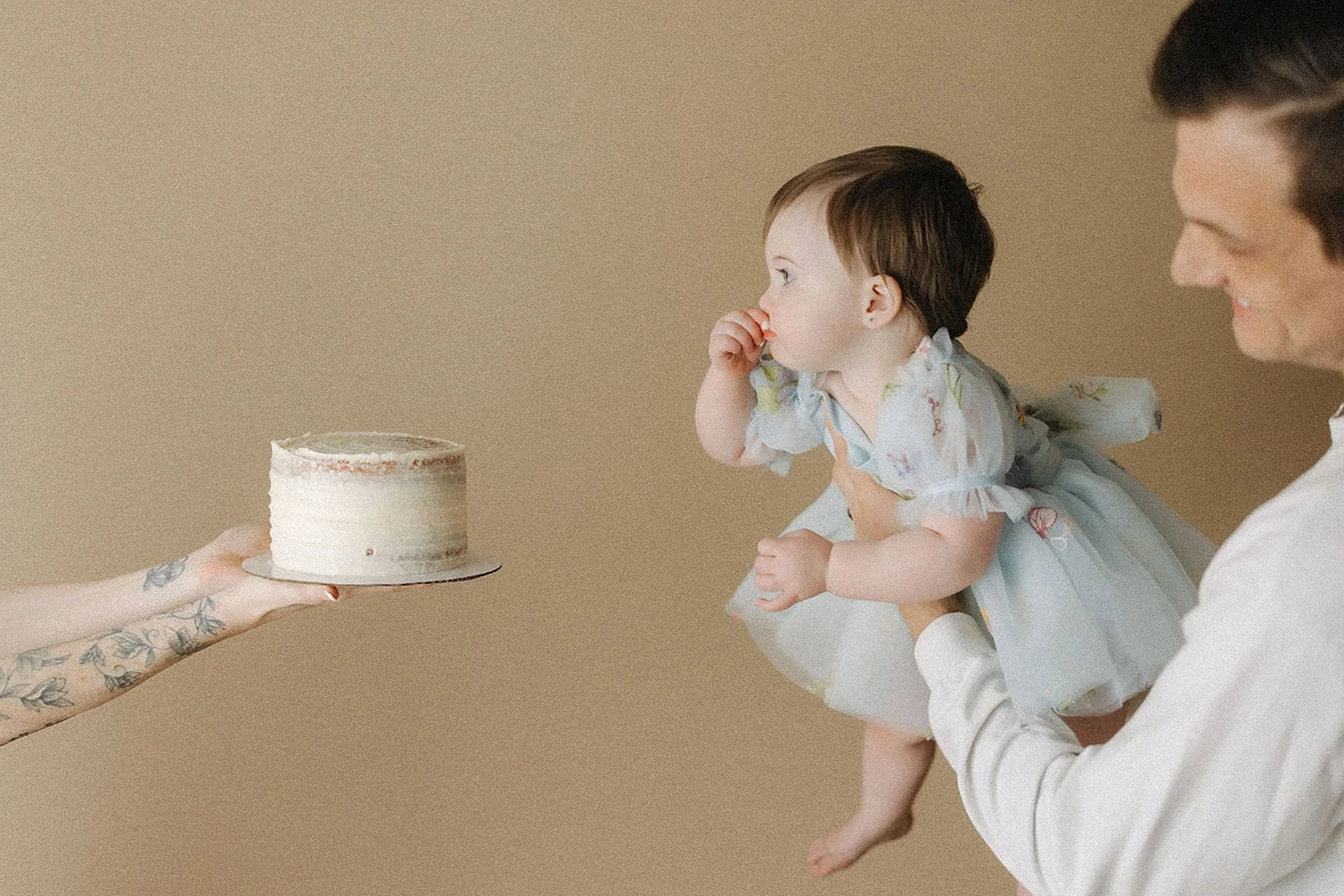 Baby girl held by her parent, reaching toward a small white buttercream cake presented by a tattooed hand, playful setup shot for a simple cake smash photography session
