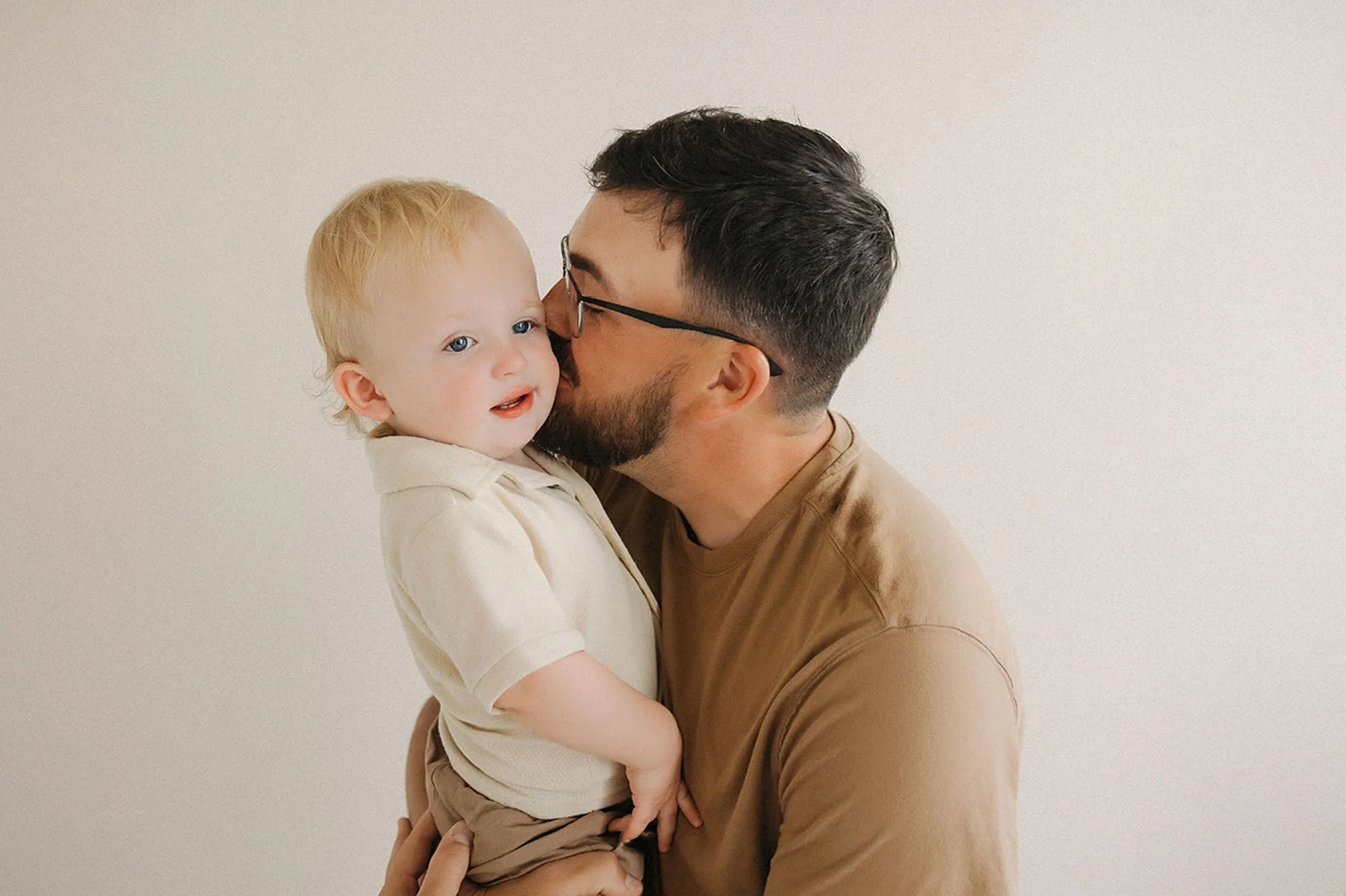 Tender studio portrait of a bearded father holding his blond toddler and kissing him gently on the cheek, both dressed in warm neutral tones against a soft cream backdrop