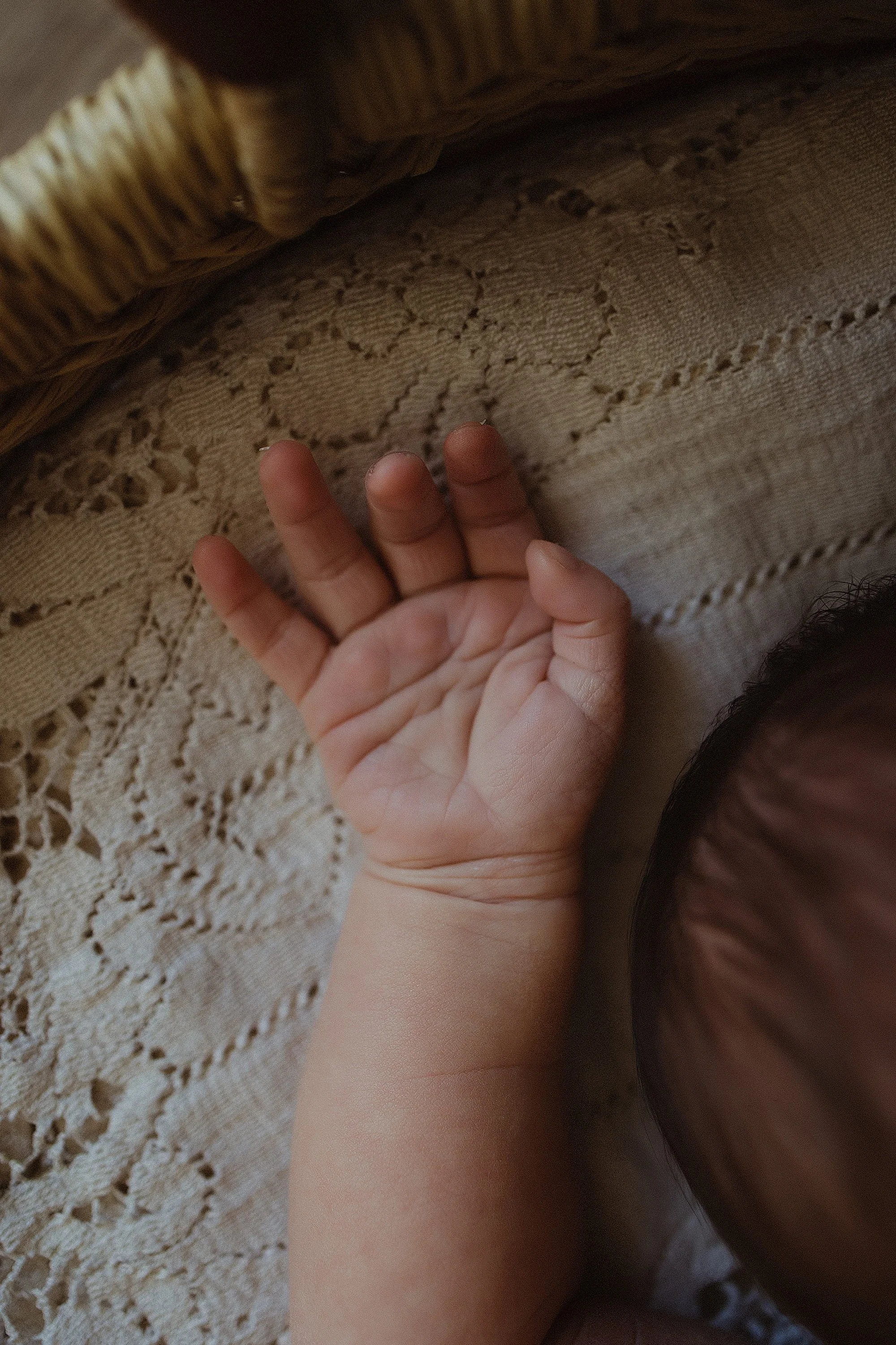 Close-up of a baby's hand lying on a lace tablecloth, with part of the baby's head visible in the lower right corner.