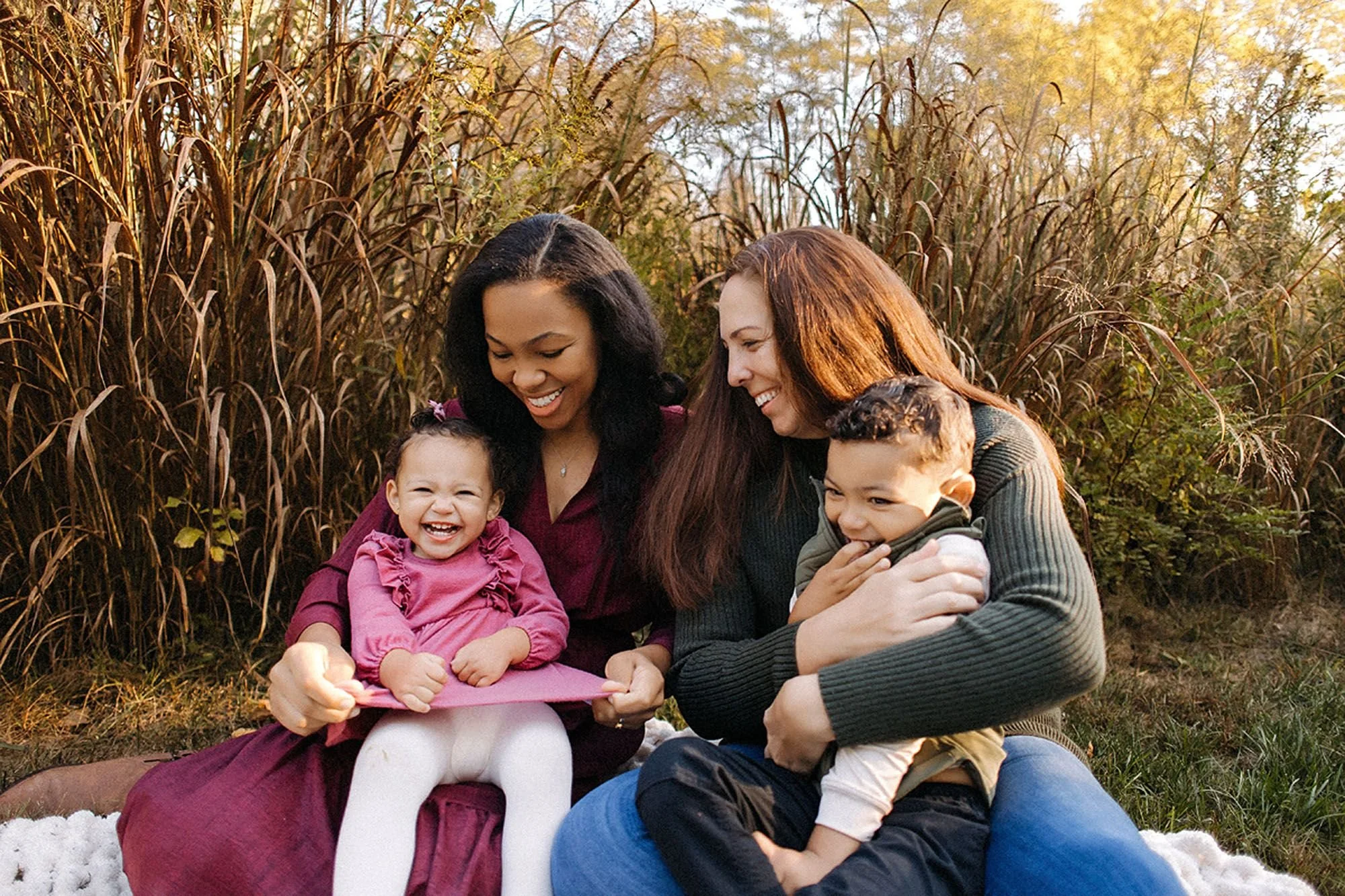 Two mothers laughing with their toddlers during a family photoshoot in tall autumn grasses at Virginia B. Fairbanks Art & Nature Park, capturing joyful, candid moments in nature