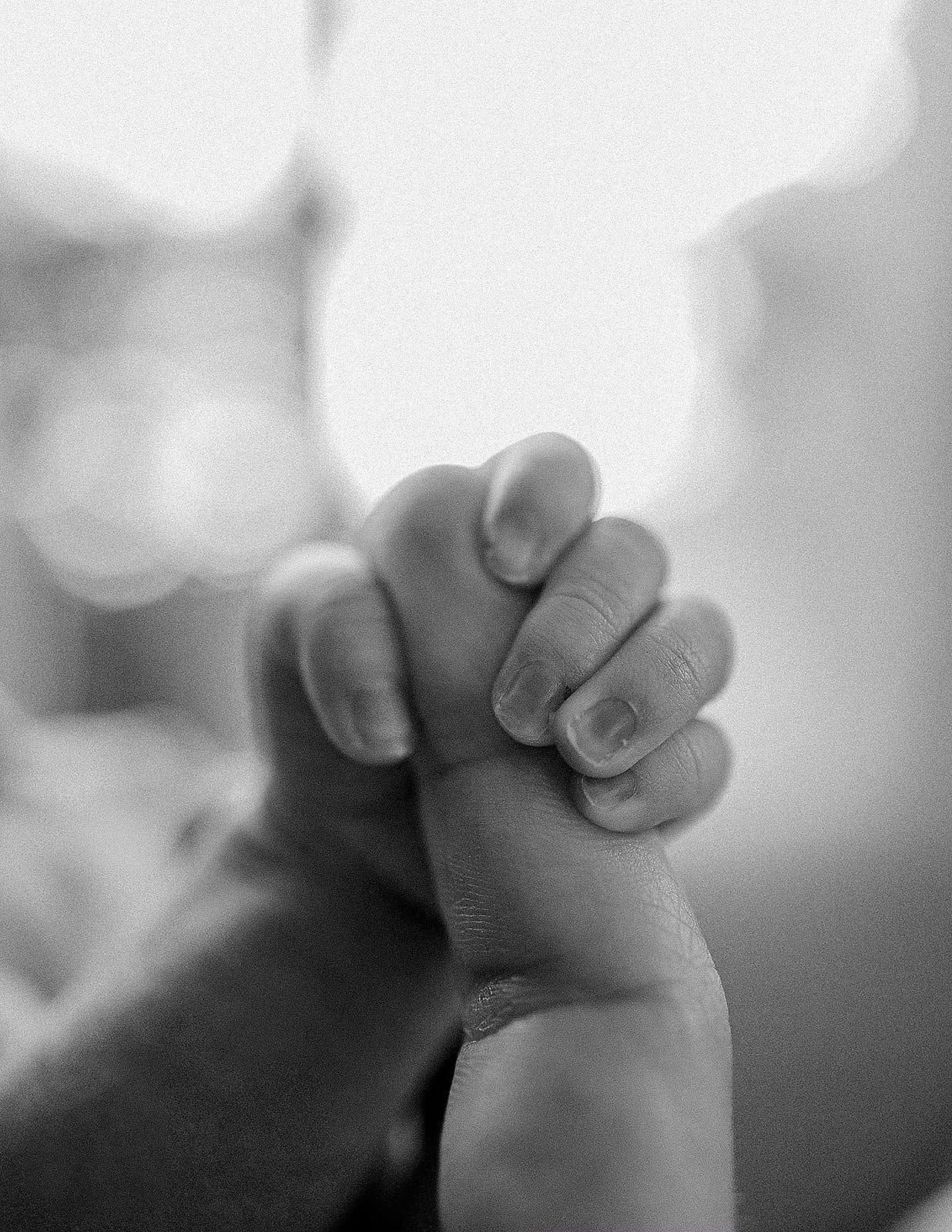A close-up black and white photograph of an adult's hand gently gripping a baby's hand.