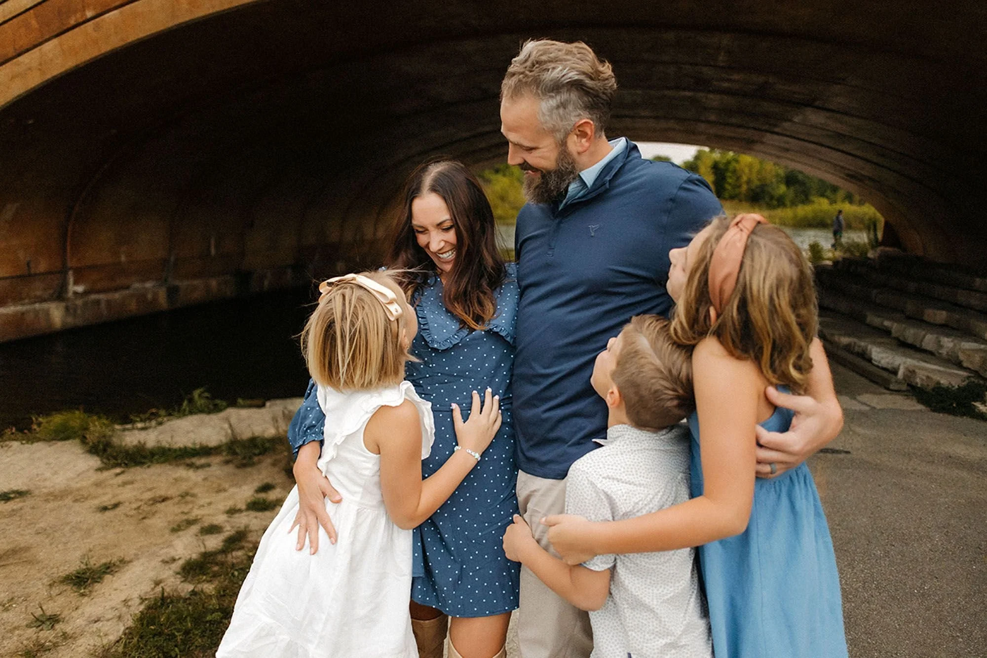 Parents and children sharing a joyful group hug beneath a scenic bridge during a warm outdoor family photography session in Carmel, Indiana