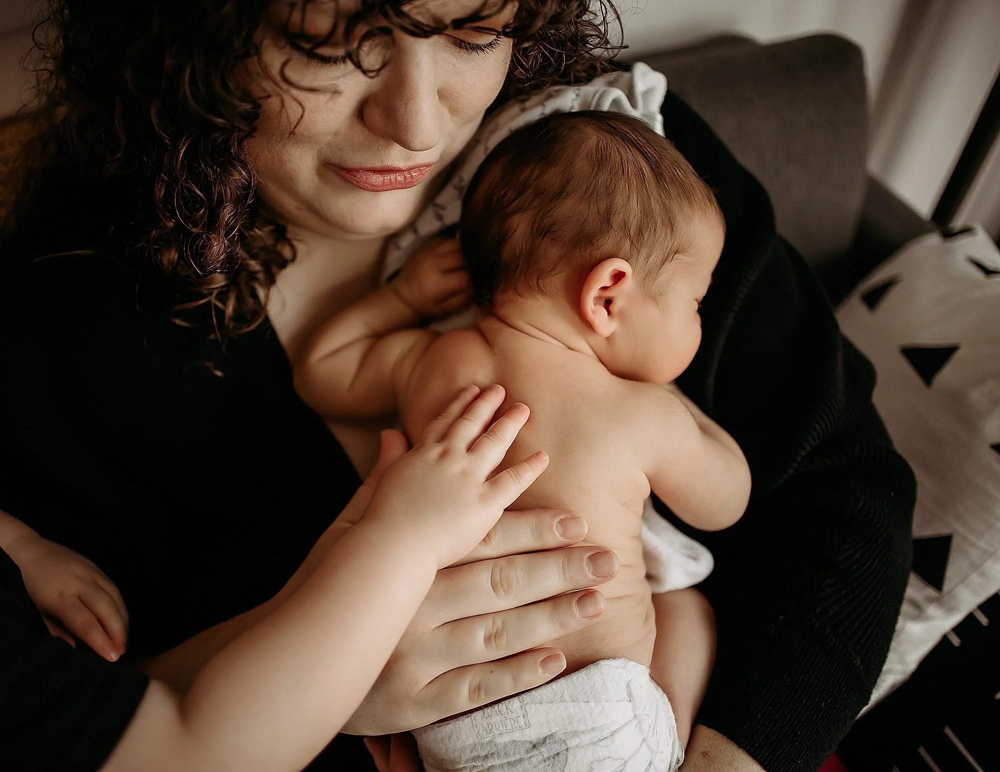 Mother holds newborn baby doing skin to skin while toddler sibling pats the newborn's back during indianapolis newborn photography photoshoot
