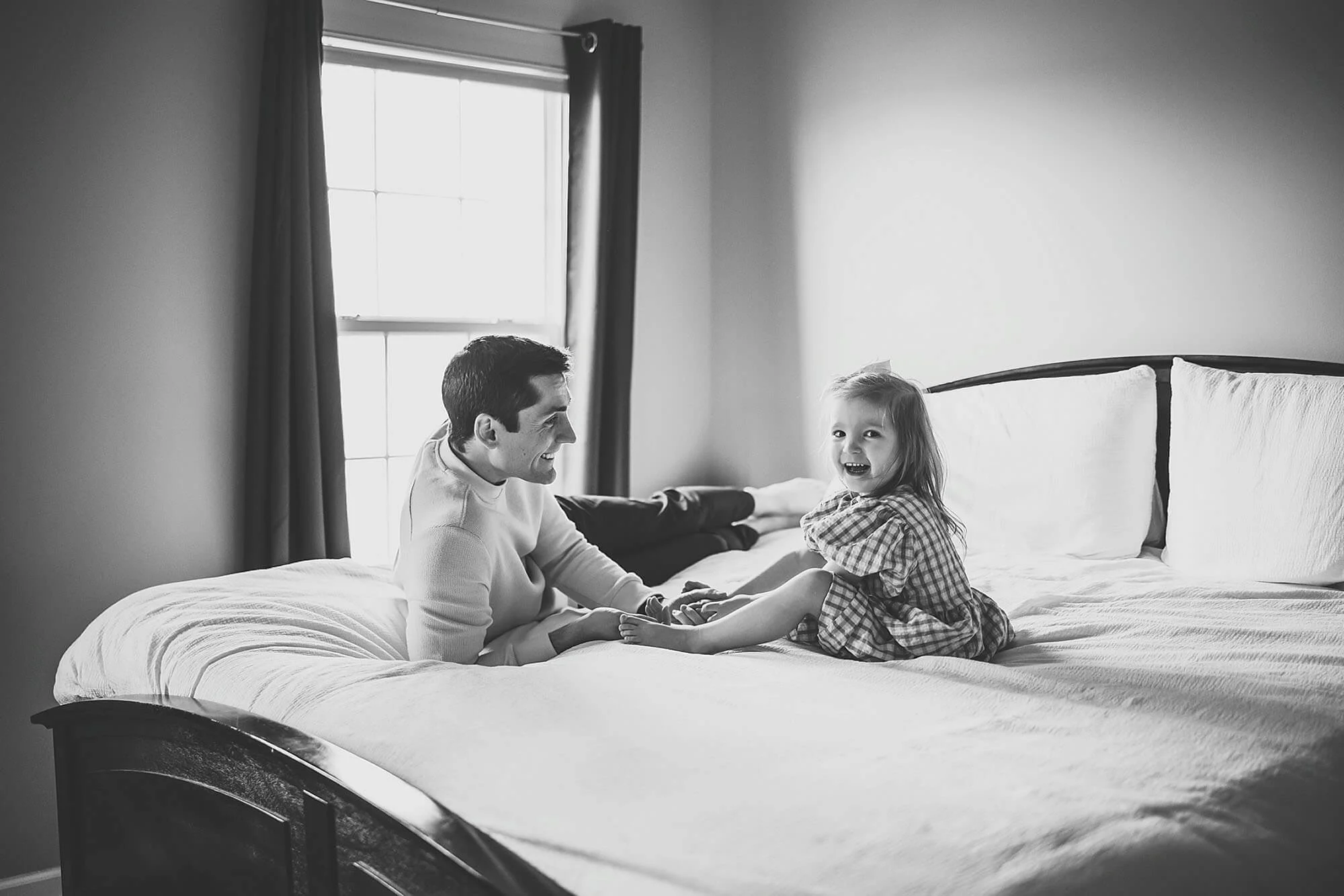 A father and a little girl are seated on a bed, enjoying a moment during their New Baby Photos session